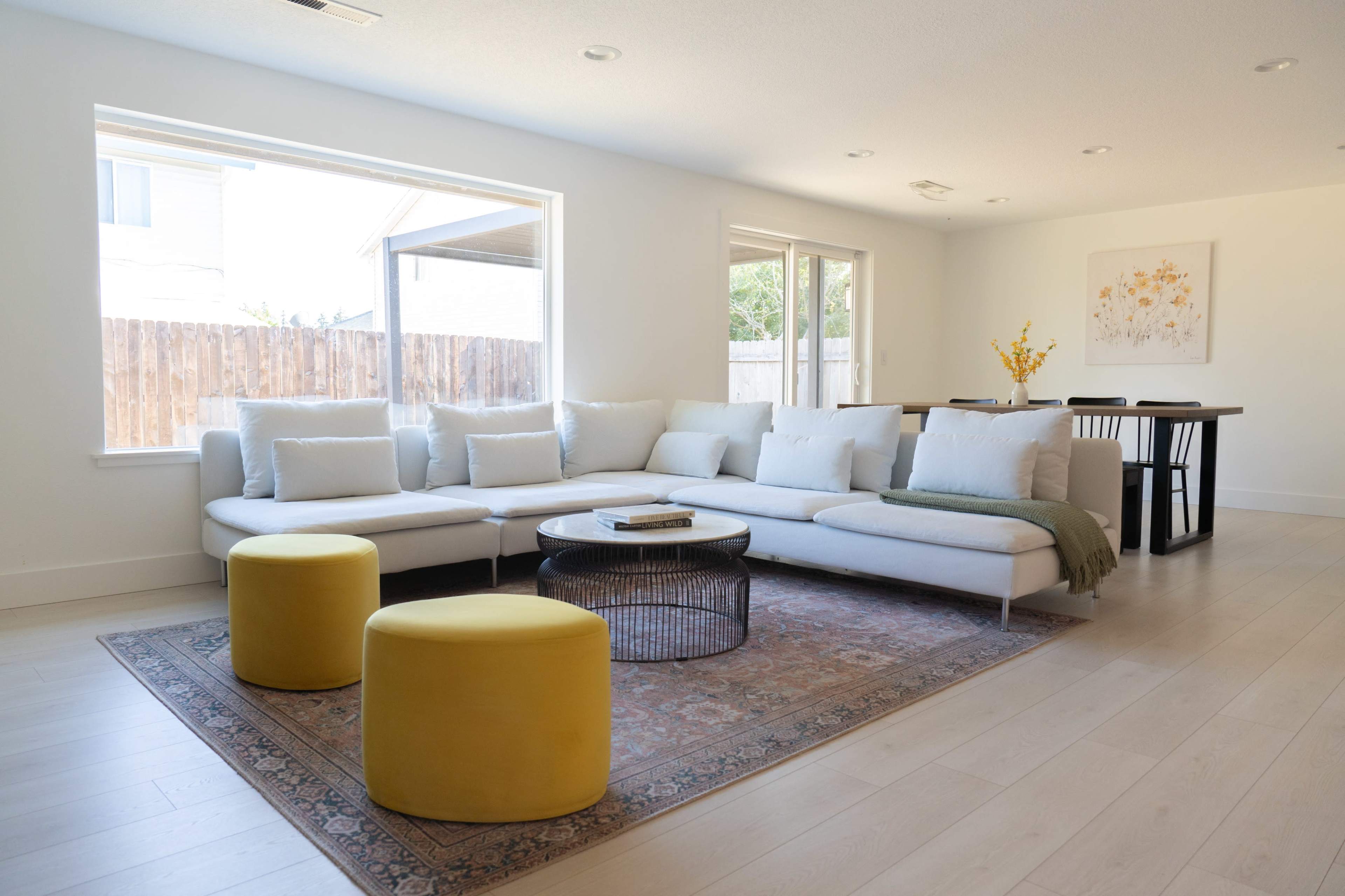 A light-filled living room features a sectional sofa with pillows, two yellow ottomans, a round coffee table, and a dining area in the background.