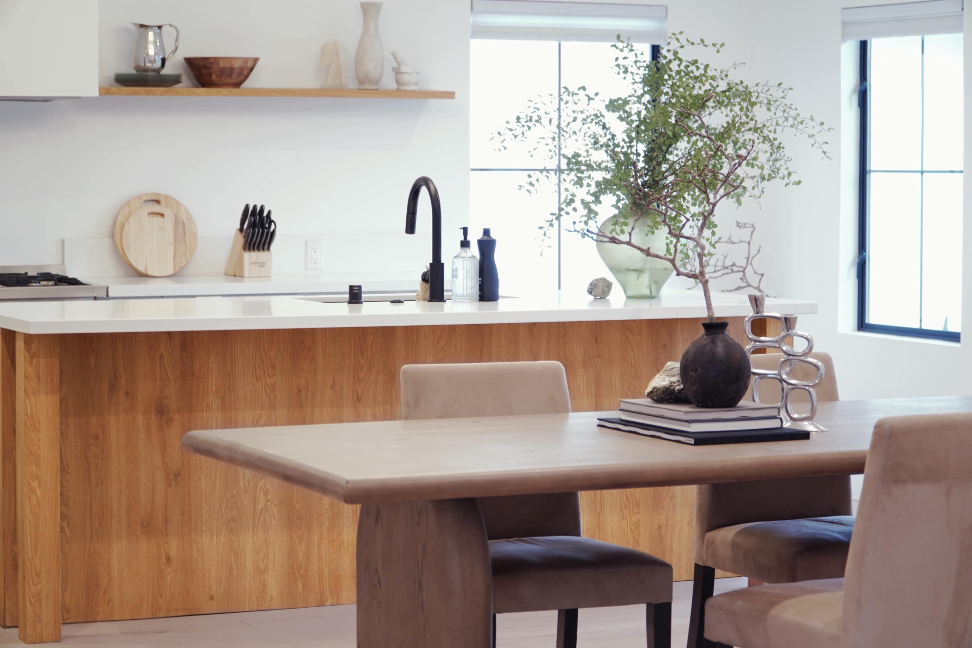 A modern kitchen and dining area features wooden furnishings, a minimalist design, and a decorative plant centerpiece on the table.