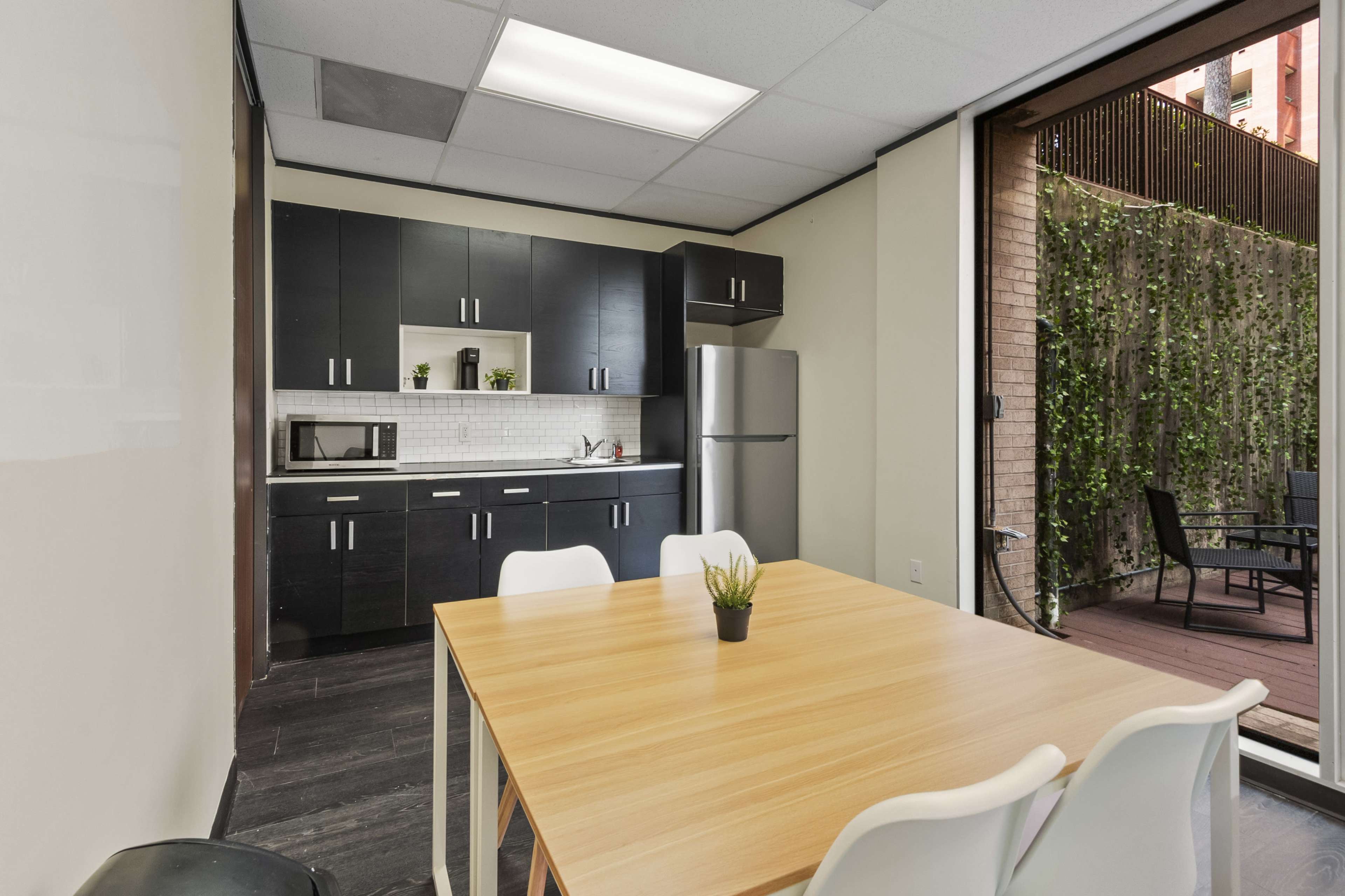 The image shows a modern kitchen and dining area with a wooden table, white chairs, and a kitchenette featuring dark cabinets, a refrigerator, and a microwave.
