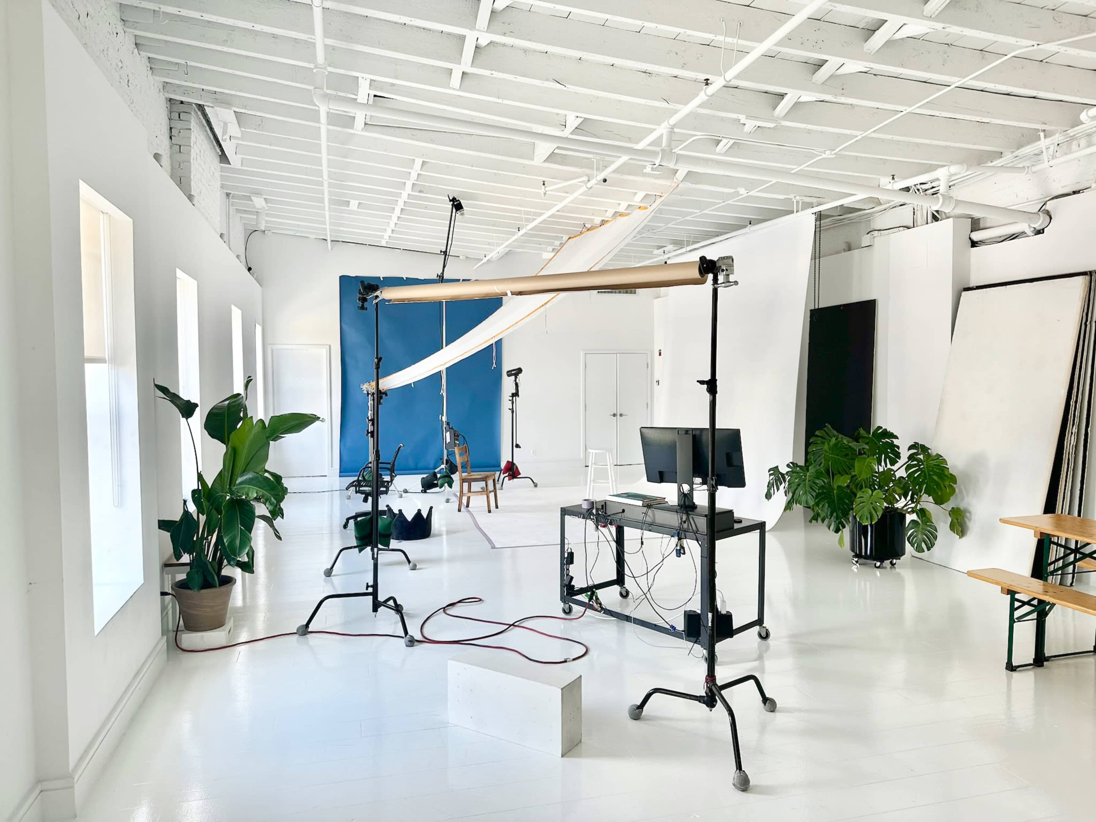The image shows a bright studio space with white walls, a long table, potted plants, and equipment for photography, including backdrop supports and a computer setup.
