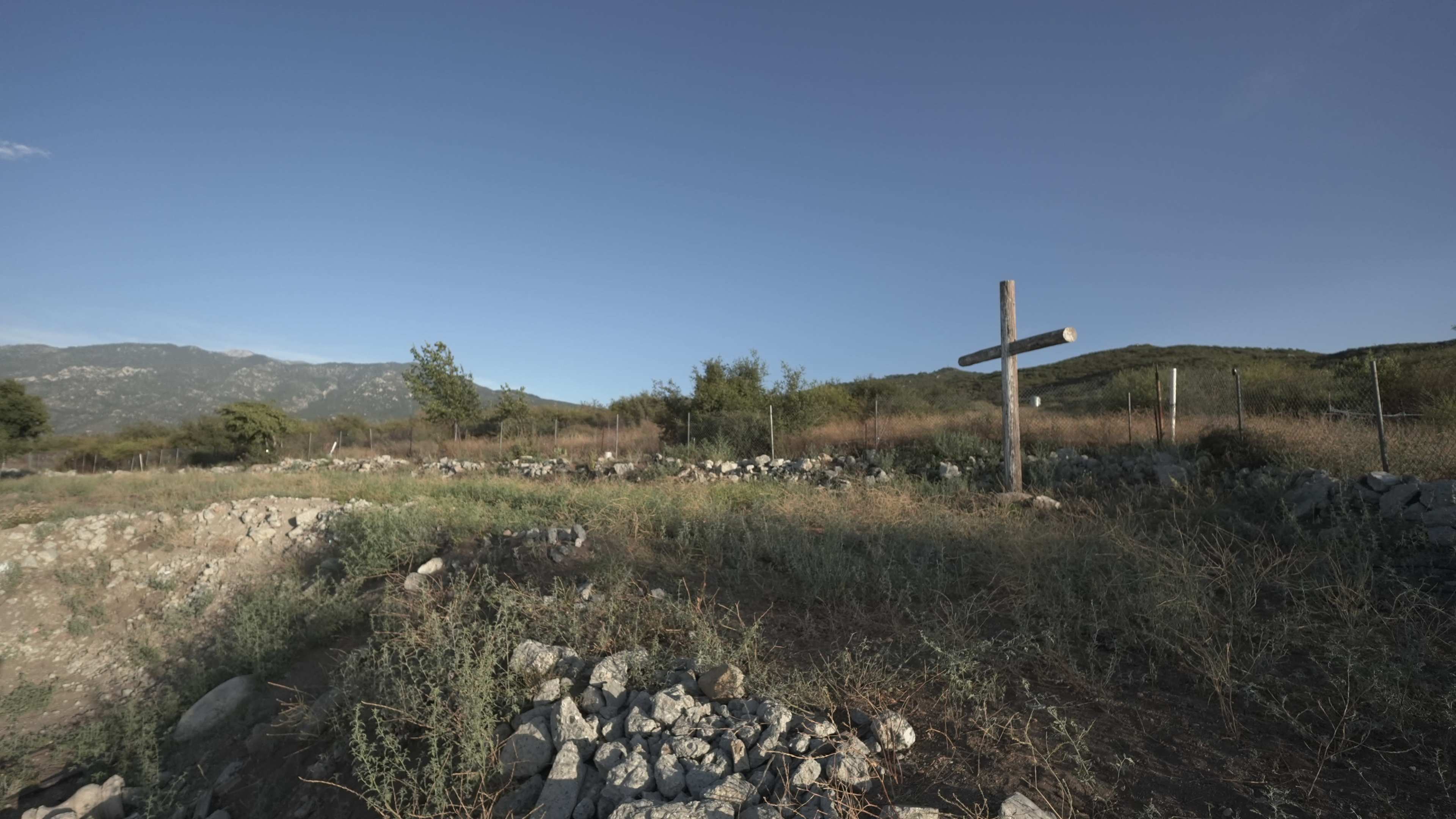A wooden cross stands among rocky terrain and sparse vegetation under a clear blue sky.