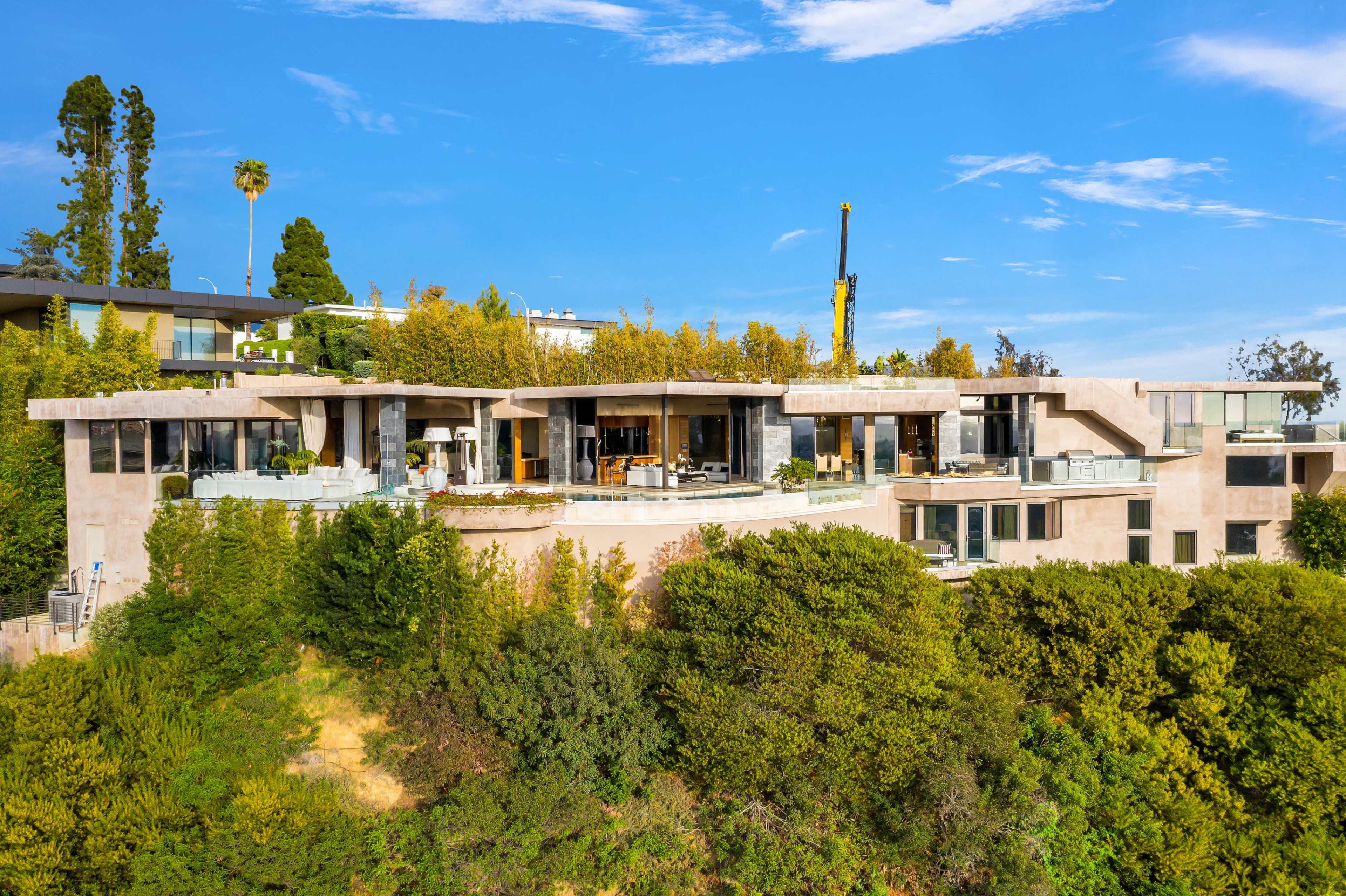 A modern multi-level house situated on a hillside, surrounded by greenery and featuring large glass windows and a patio area.