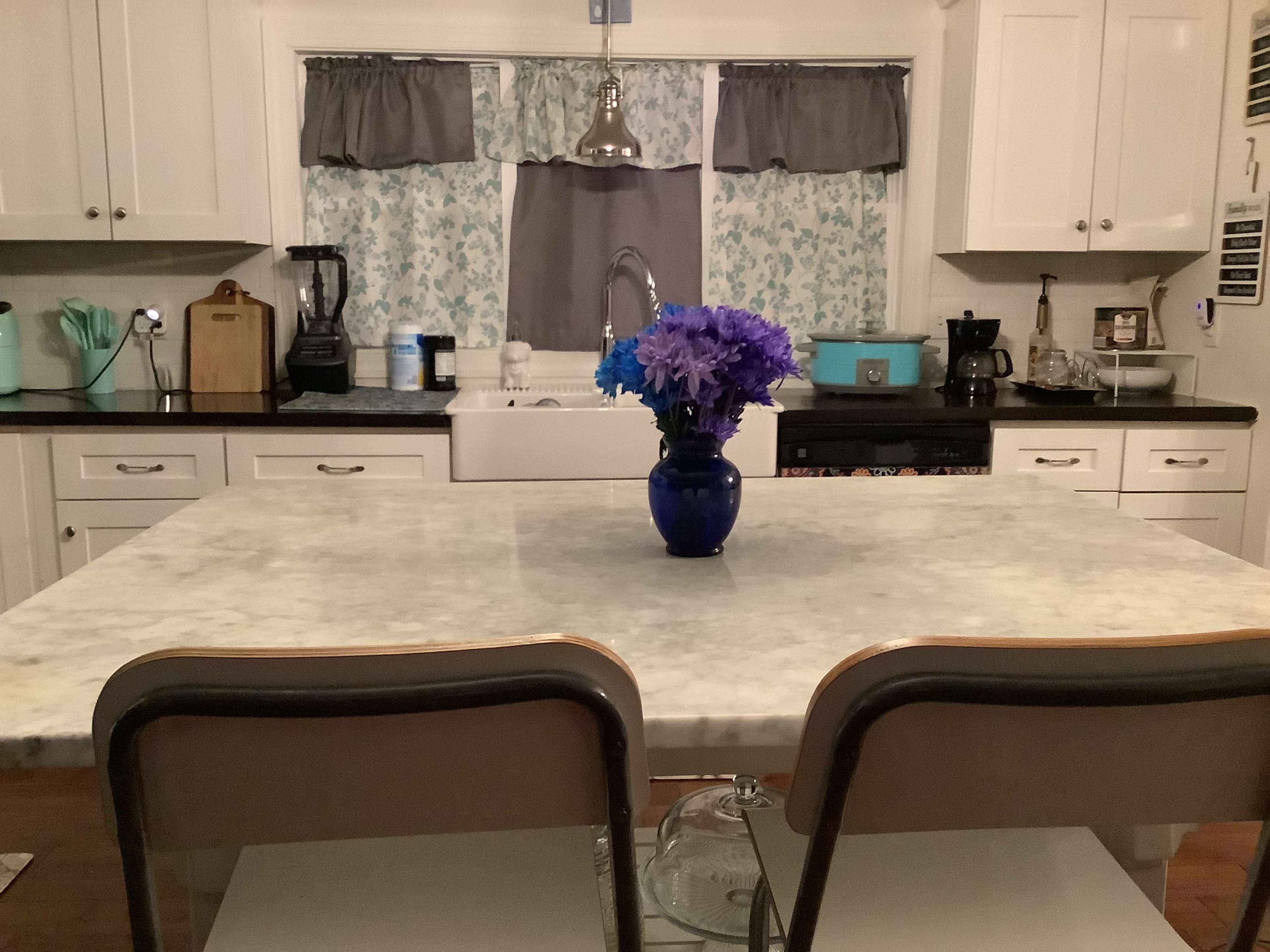 A kitchen features a marble table with two chairs and a vase of flowers in the center, set against a backdrop of cabinetry and appliances.