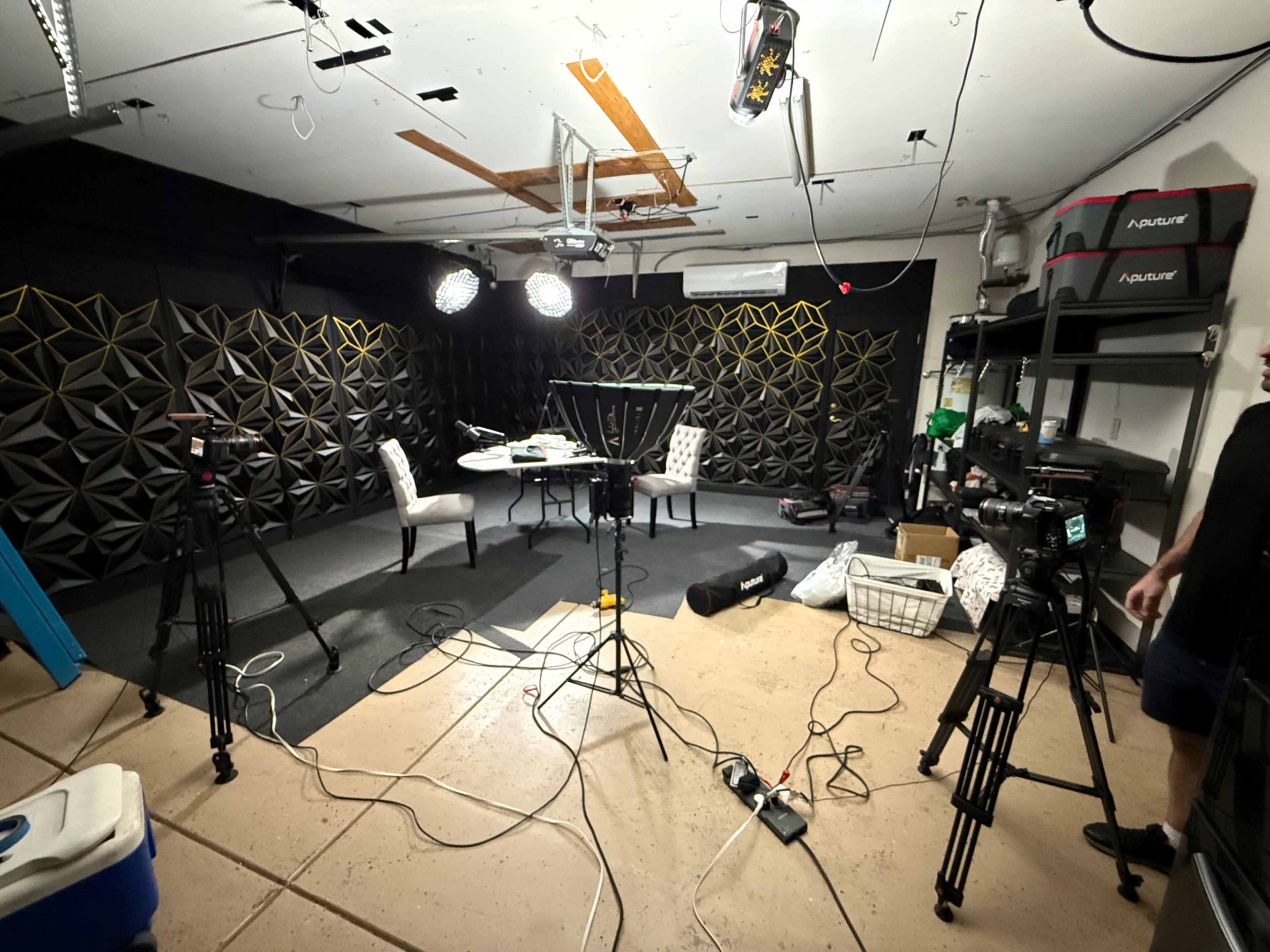 The image shows a video recording studio equipped with cameras, lighting, and a table surrounded by chairs.