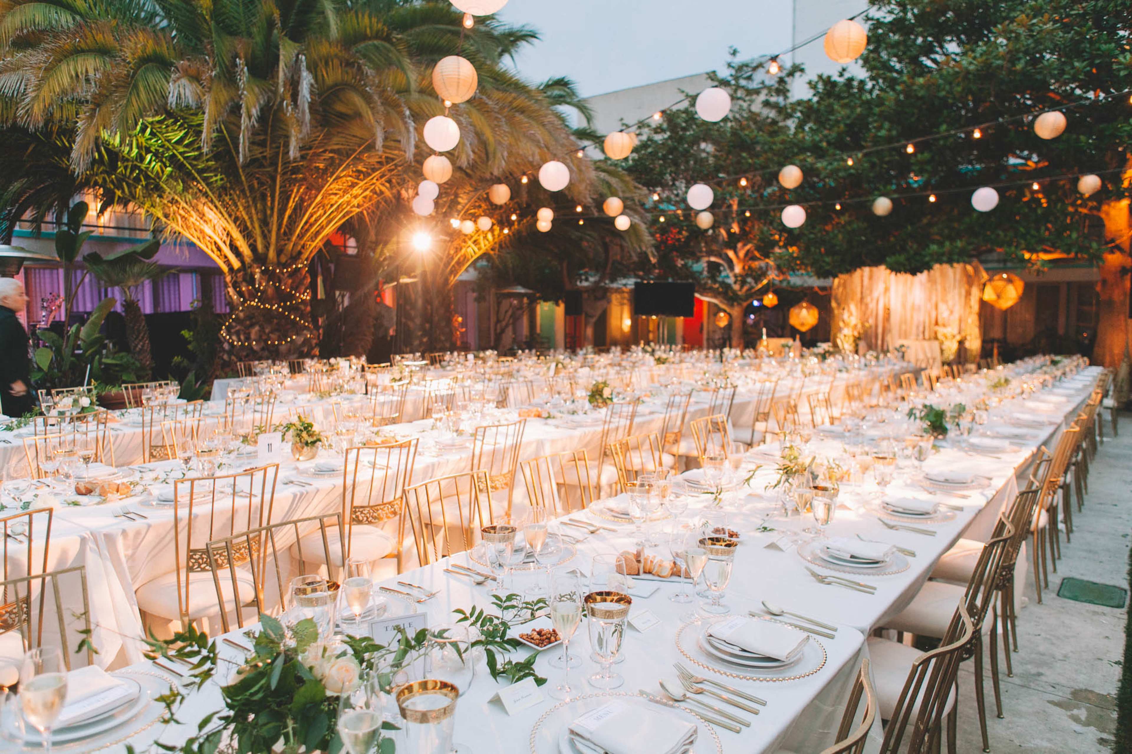 A long dining table is elegantly set under palm trees, adorned with white tablecloths, glassware, and decorative centerpieces, with string lights and lanterns illuminating the scene.