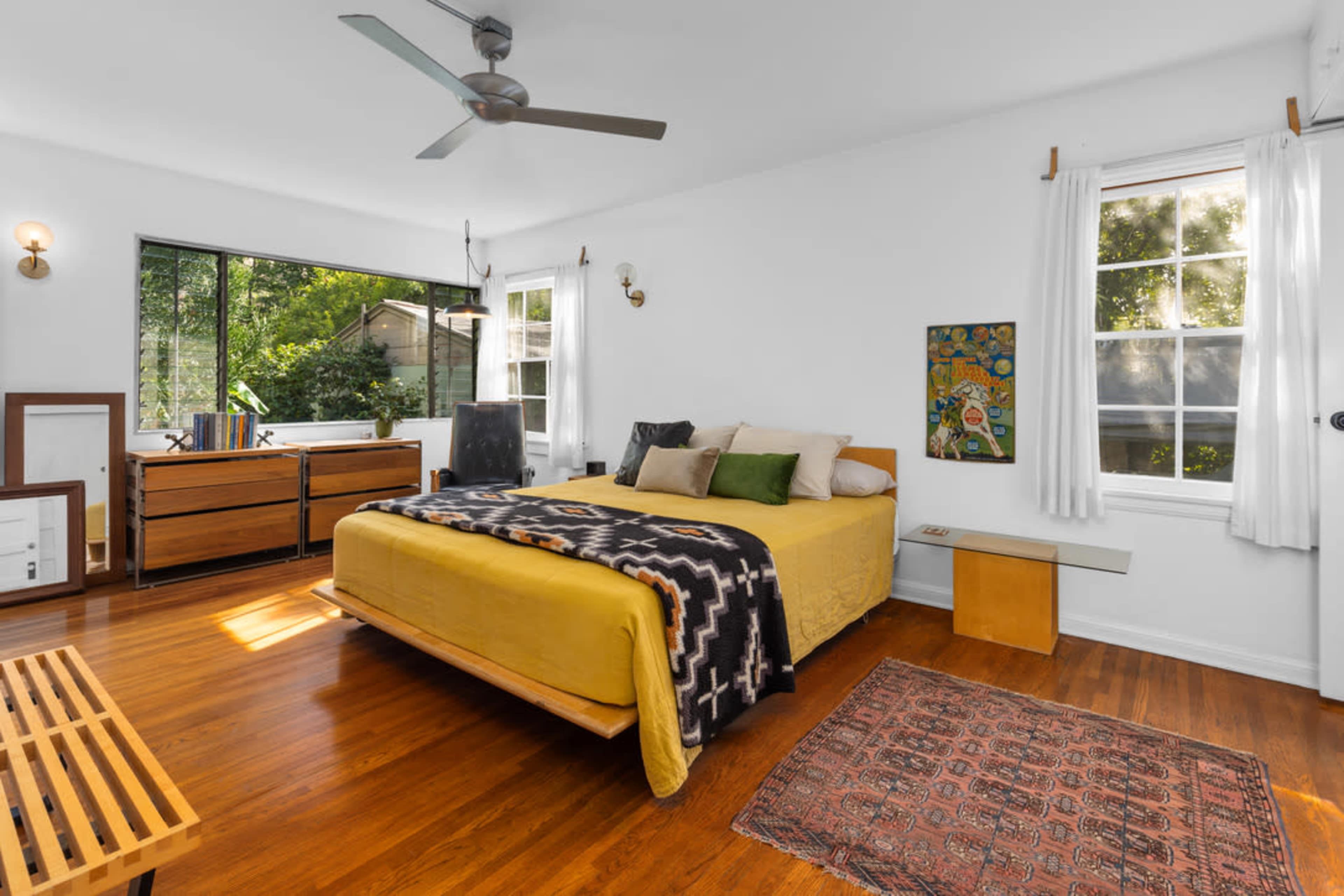 The room features a bed with a yellow coverlet, wooden furniture, and large windows that allow natural light to fill the space.