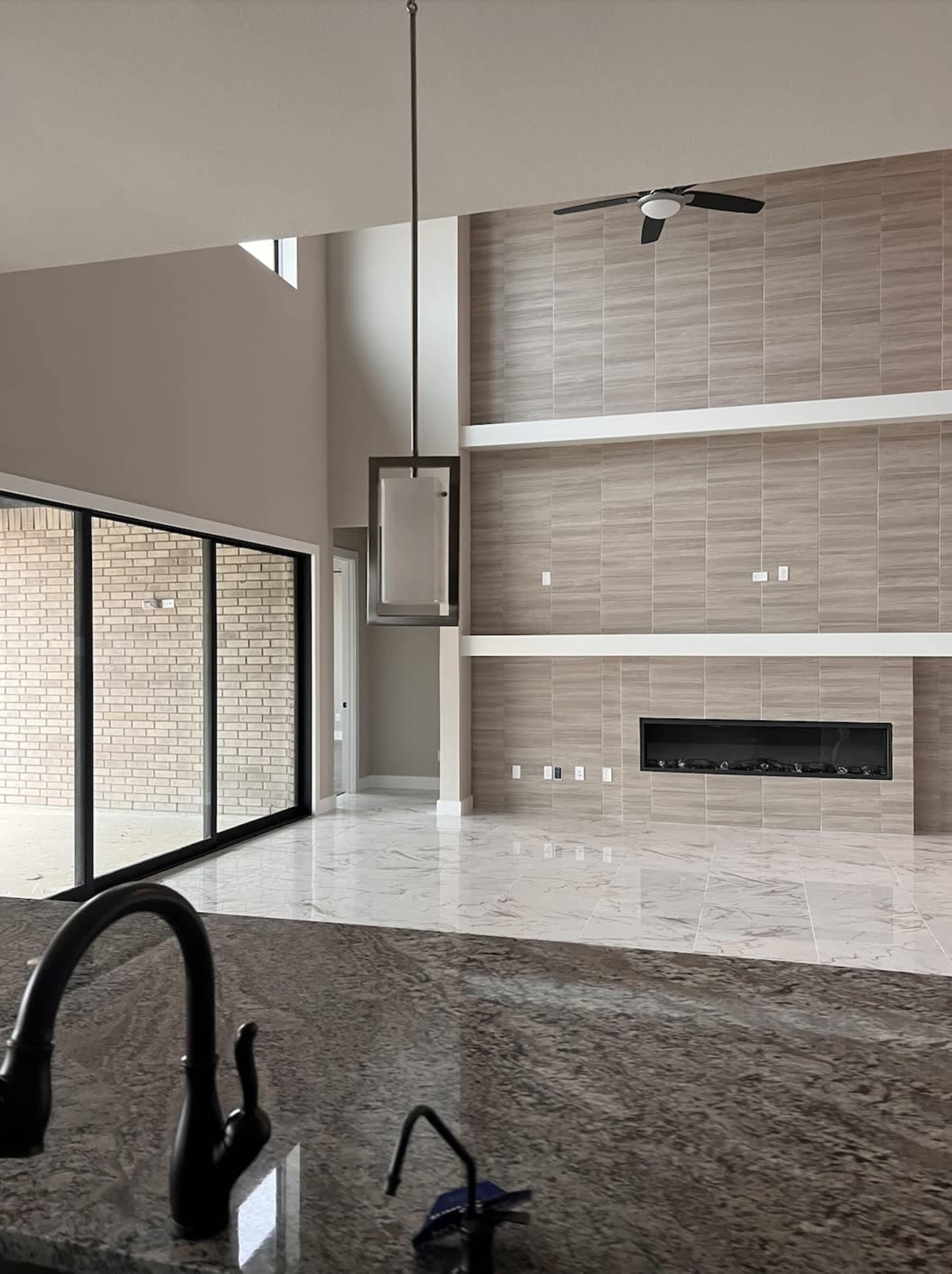 The image shows a spacious interior featuring a modern living room with a large window, tile flooring, a minimalist chandelier, and a wall-mounted fireplace.
