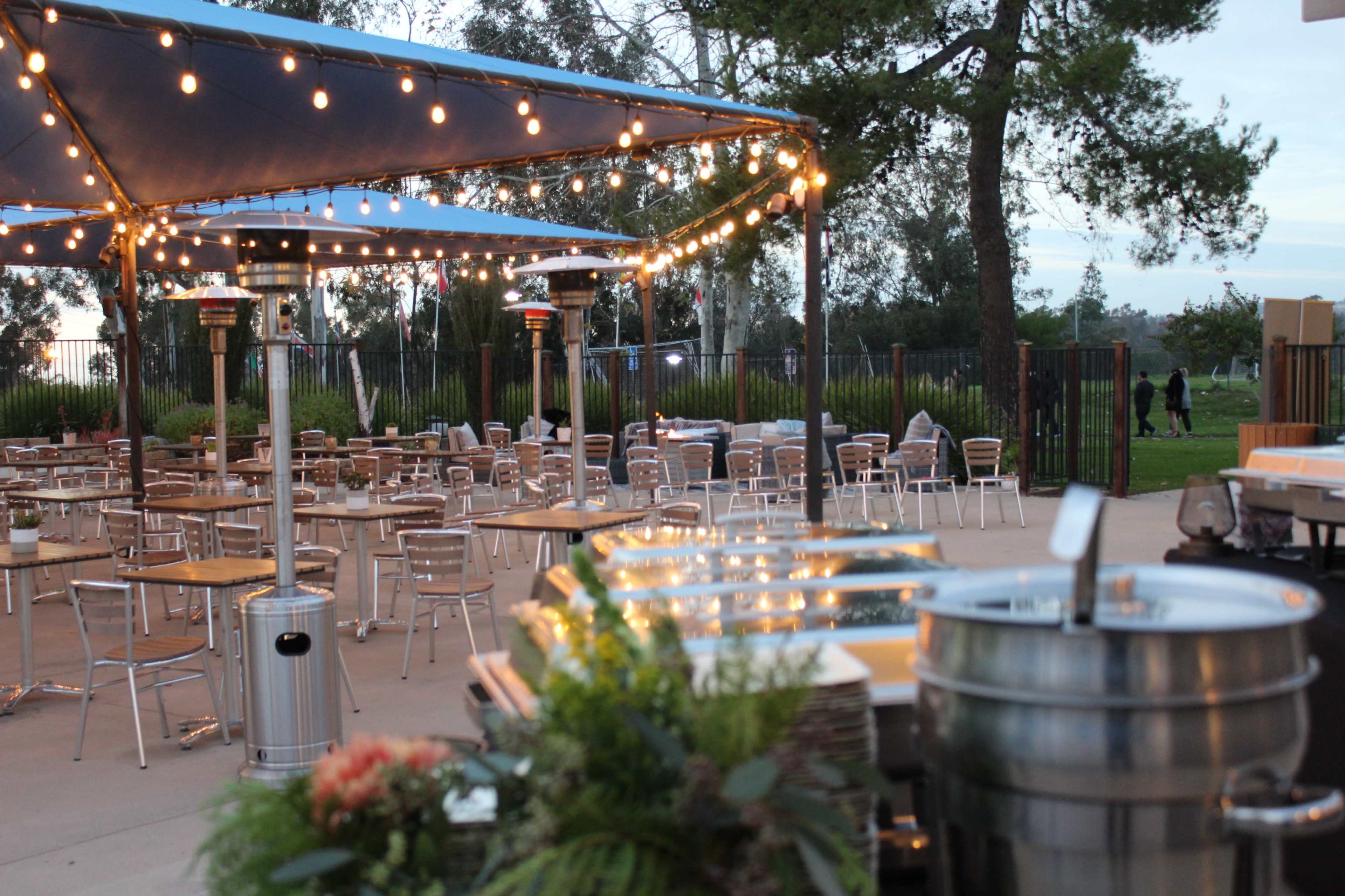 An outdoor dining area features metal tables and chairs under a canopy, illuminated by string lights, with heating lamps positioned around a central food setup.