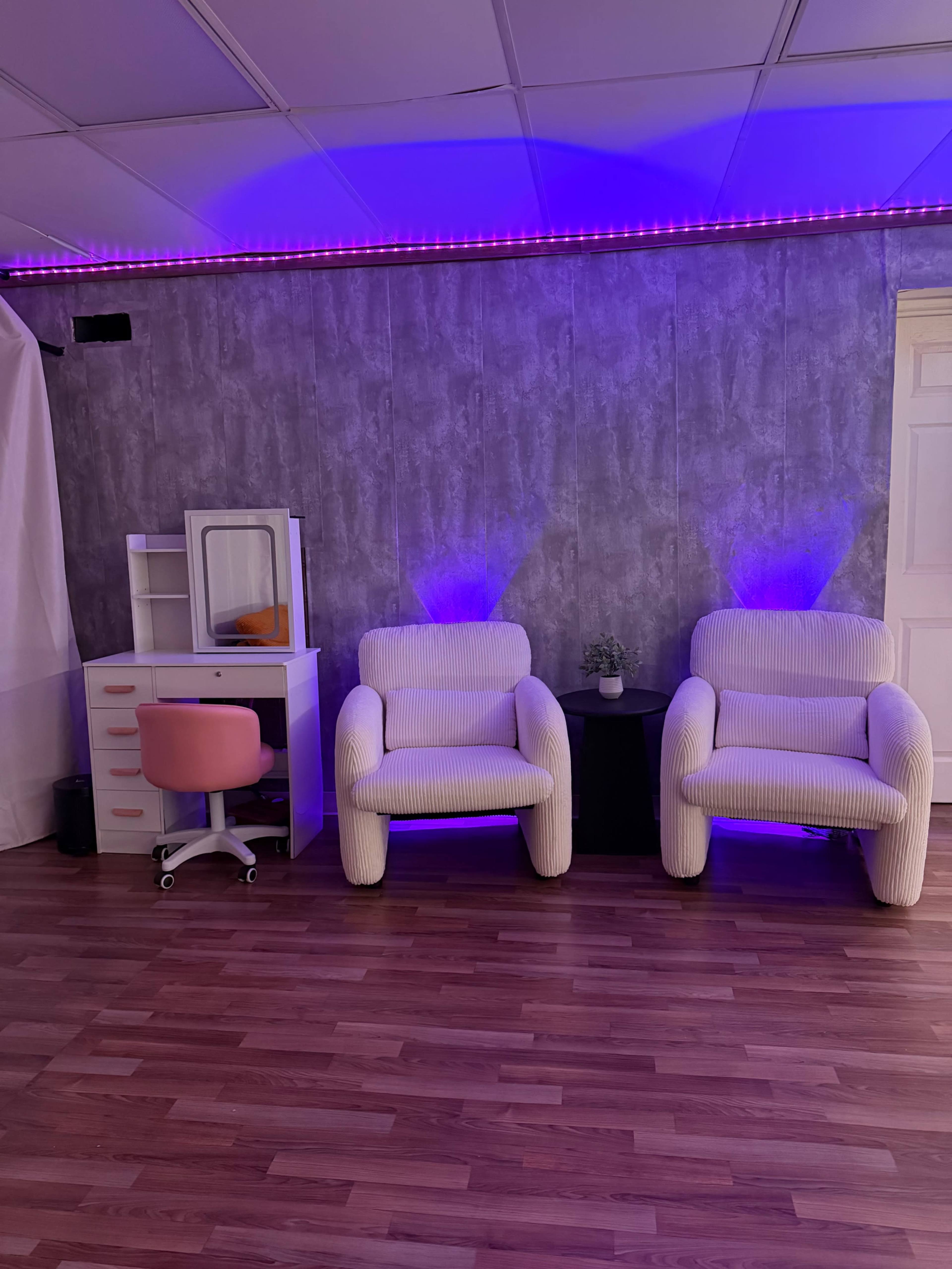 The image shows a modern space with two cream-colored chairs, a pink office chair, and a small table under soft purple lighting against a textured wall.