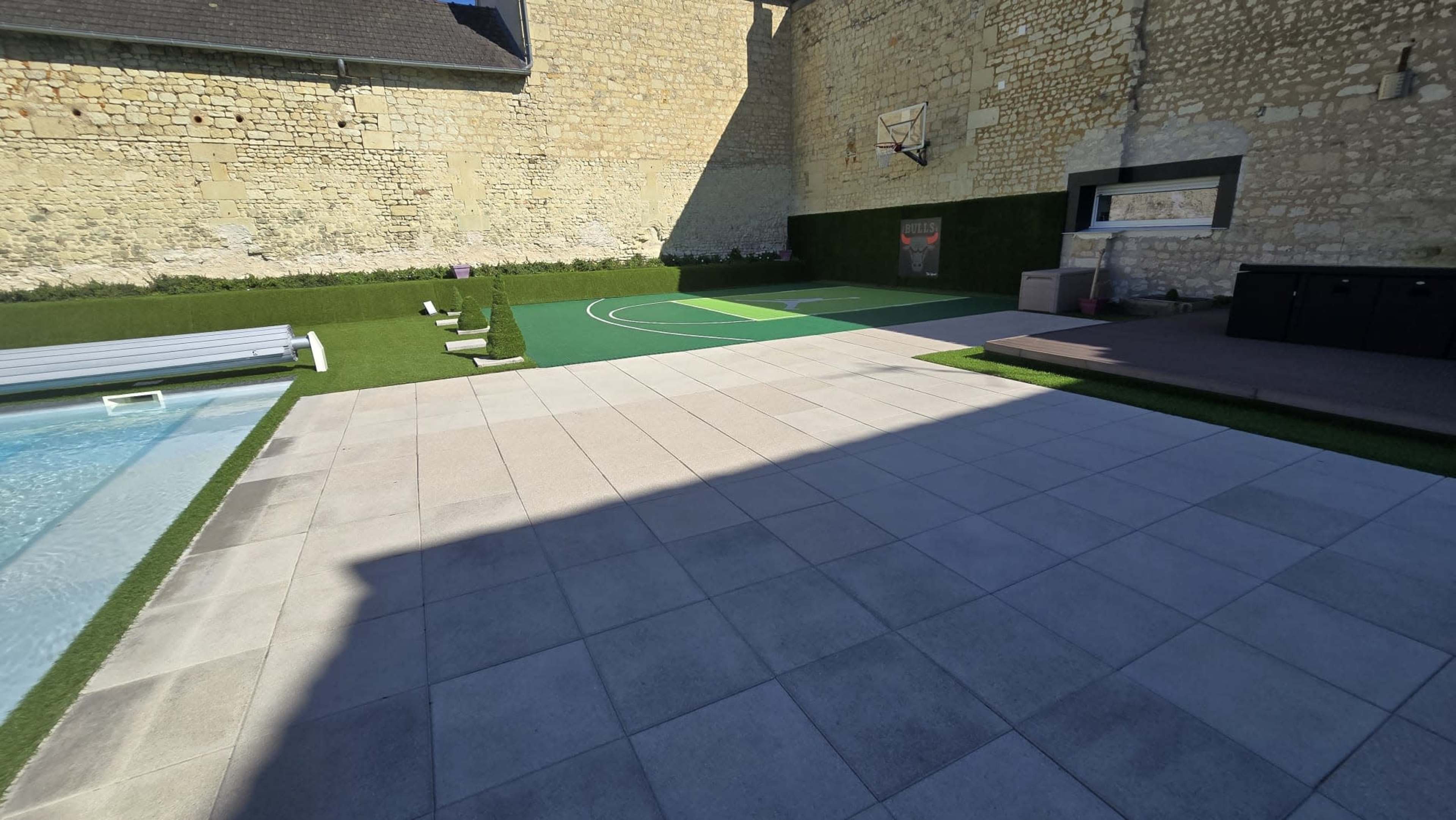The image shows a spacious outdoor area featuring a basketball court, a swimming pool, and stone flooring surrounded by grass and stone walls.