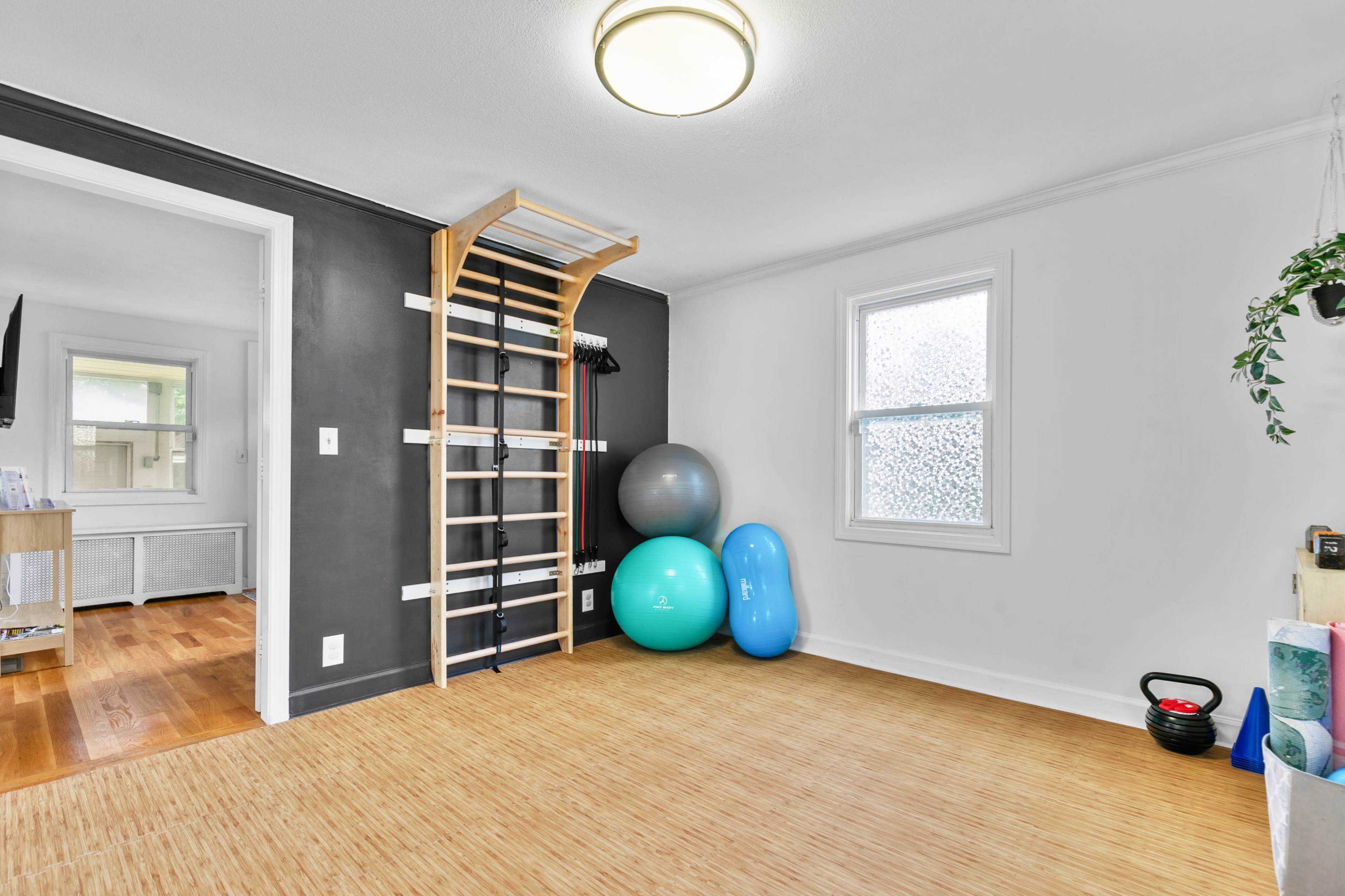 The image shows a bright exercise room with a wooden floor, a wall-mounted fitness ladder, exercise balls, and a kettlebell.