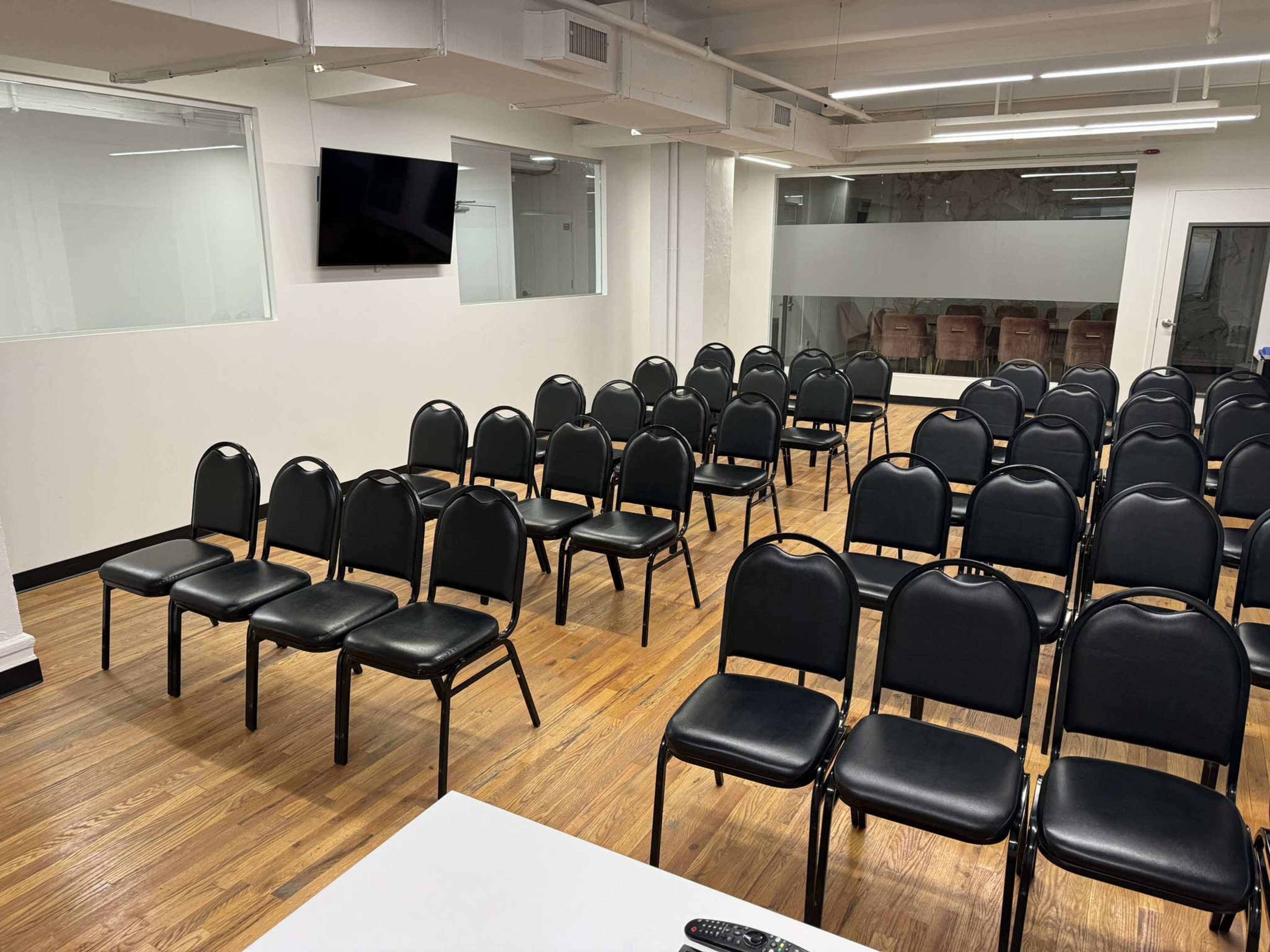 The image shows a room with rows of black chairs arranged in front of a white table and a wall-mounted television.