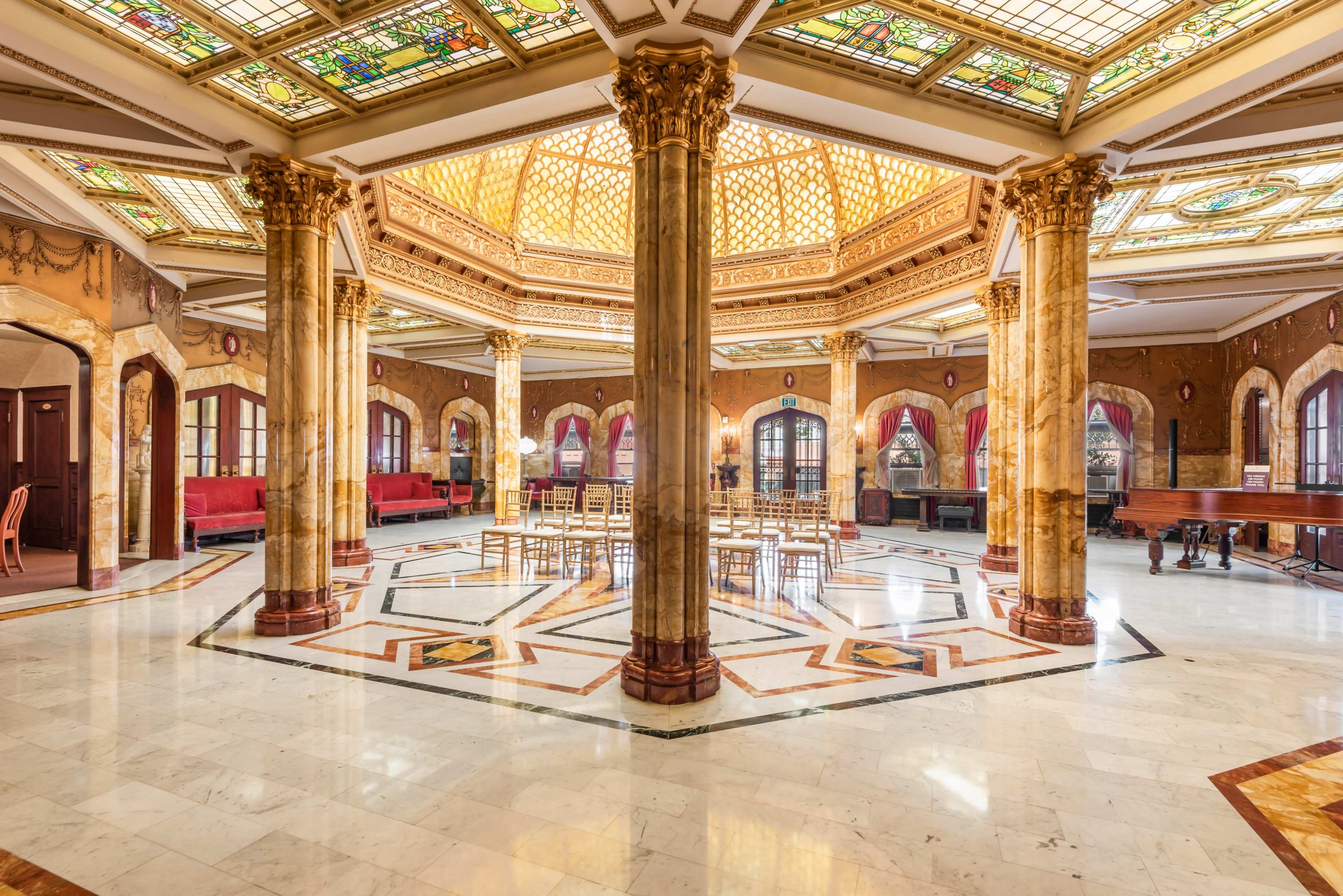 The image depicts an ornate, spacious interior with a high ceiling featuring decorative stained glass, surrounded by marble columns and elegant furnishings.
