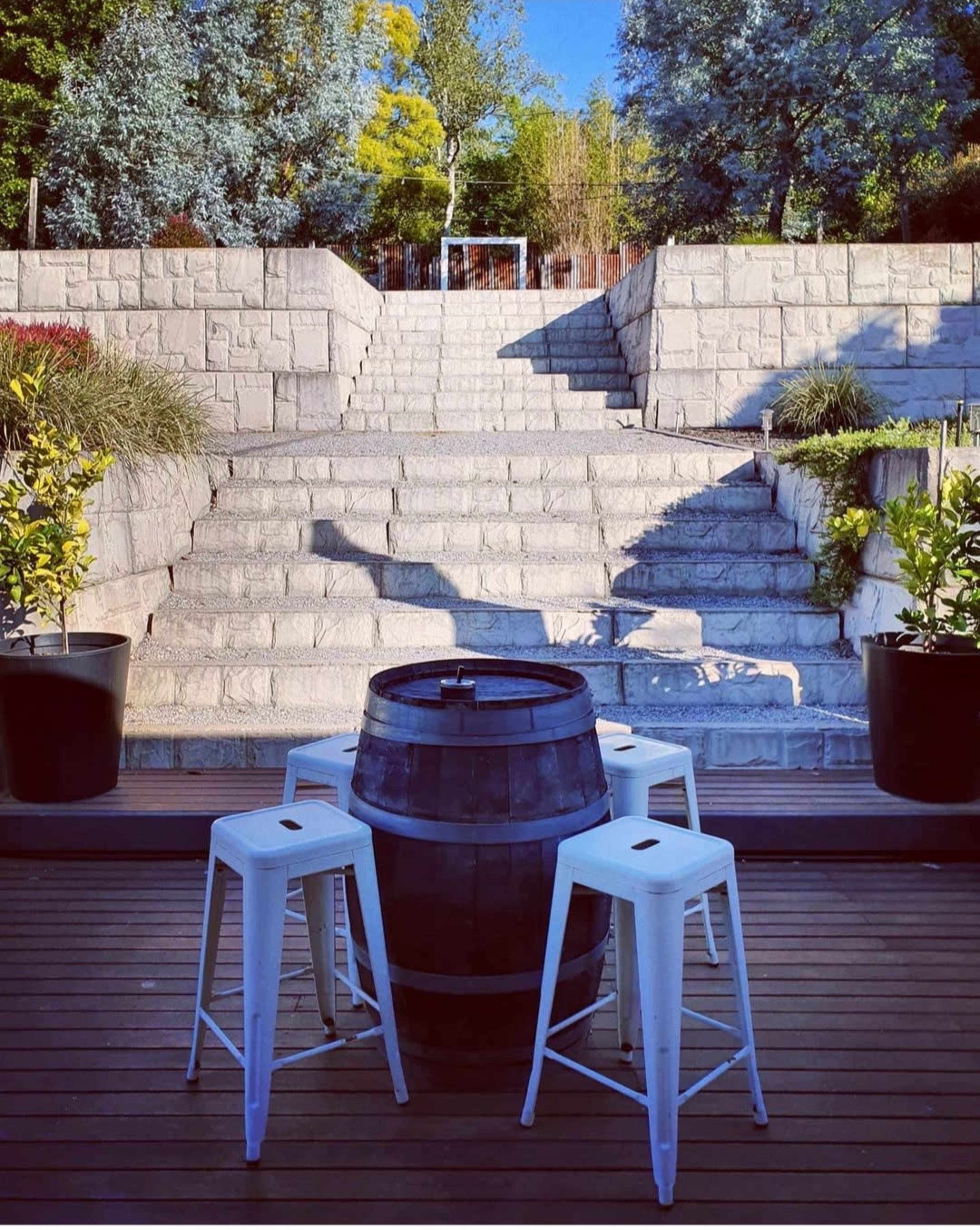 A wooden barrel stands in the center of a patio surrounded by four white stools, with stone steps leading to a landscaped area in the background.
