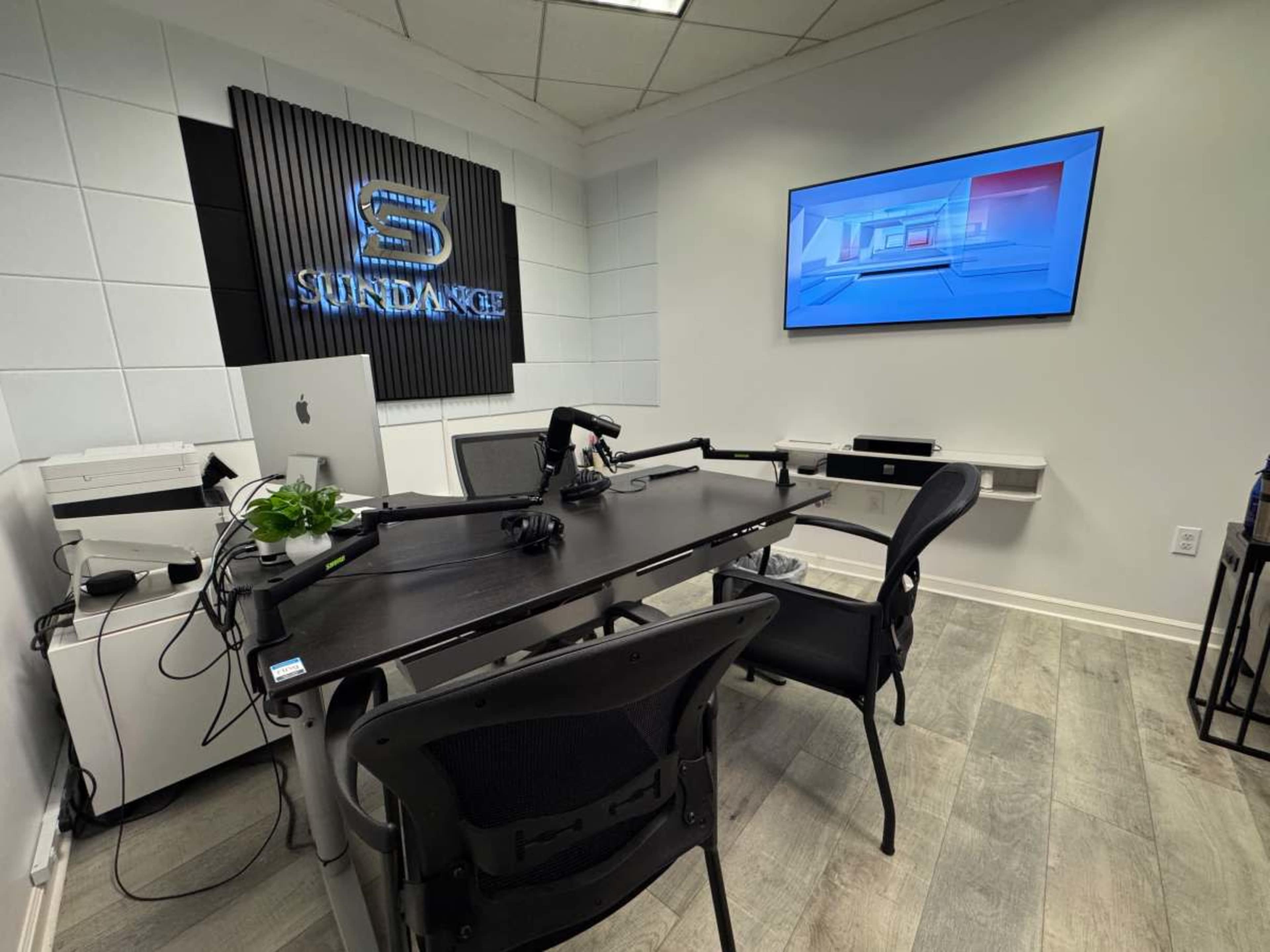 The image shows a modern office space with a black table and chairs, equipped with a microphone setup and a large screen displaying graphics on the wall.