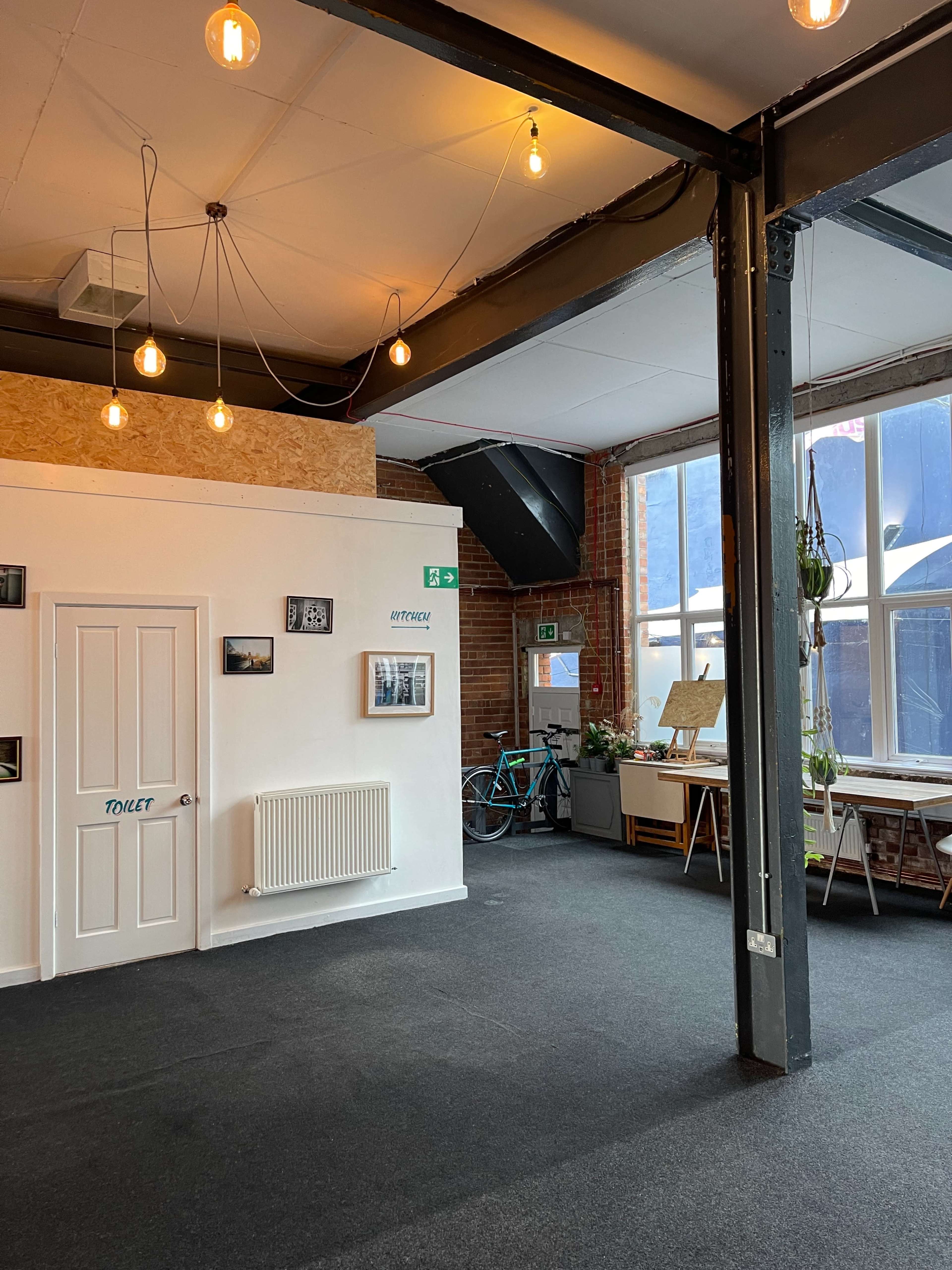 An interior space with exposed brick walls, a door labeled "TOILET," and various framed pictures hanging on the walls, alongside a bicycle and art supplies in the background.