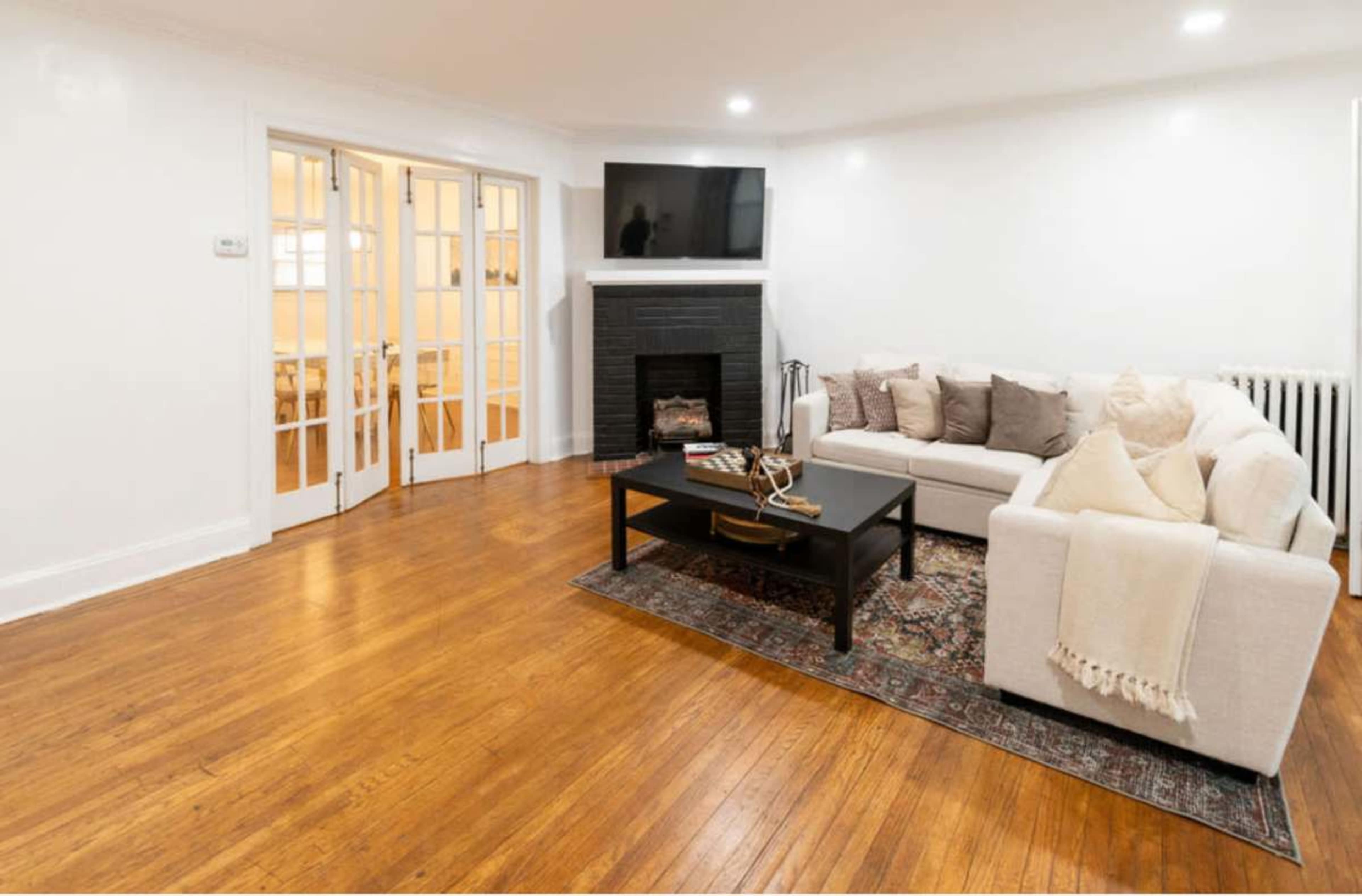 The image shows a living room with a sectional couch, a coffee table, and a fireplace, featuring hardwood flooring and a television mounted on the wall.