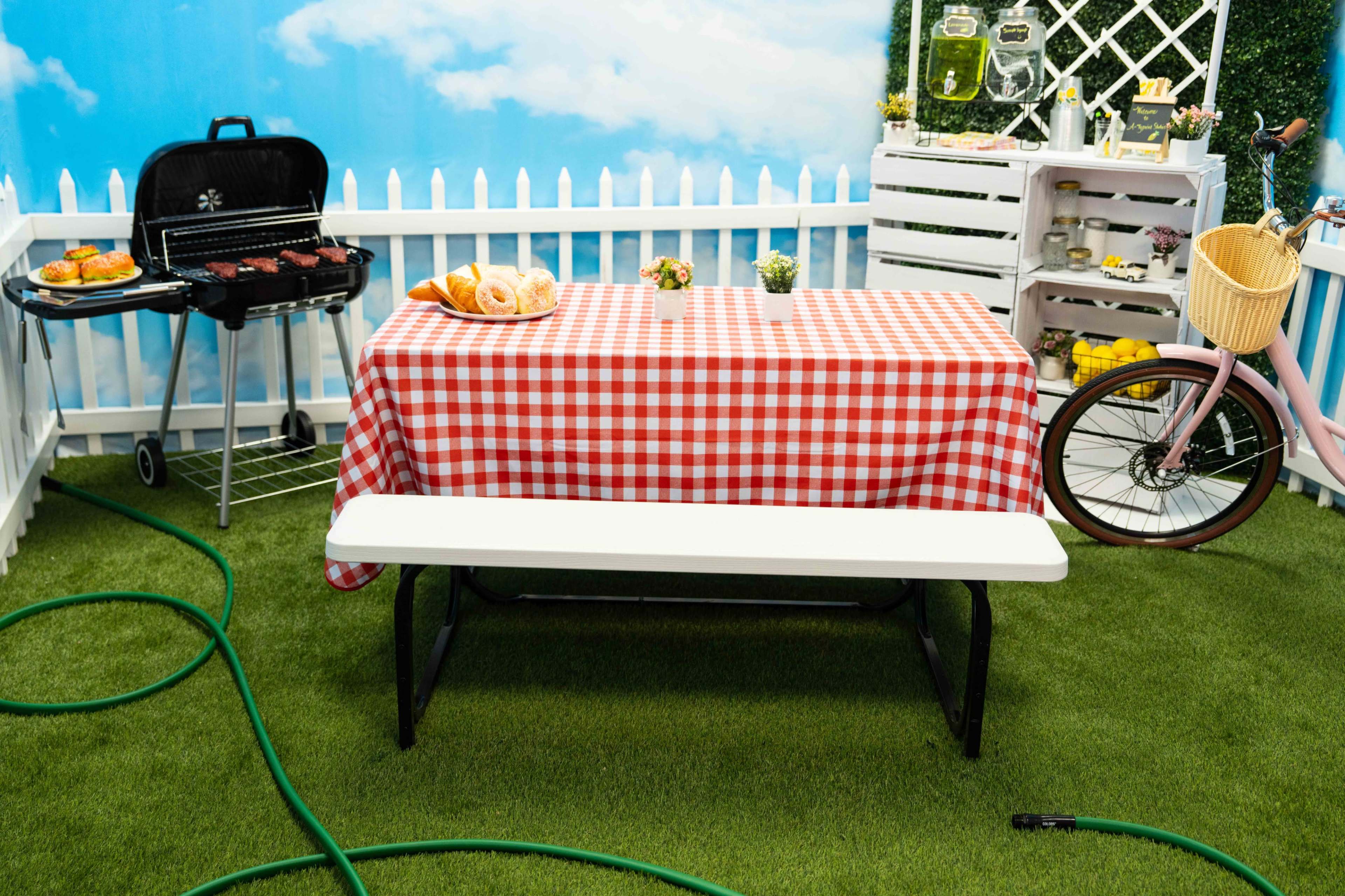 A picnic table covered with a red and white checked tablecloth is set up in a backyard, alongside a barbecue grill, a bicycle, and decorative elements against a backdrop of a blue sky.
