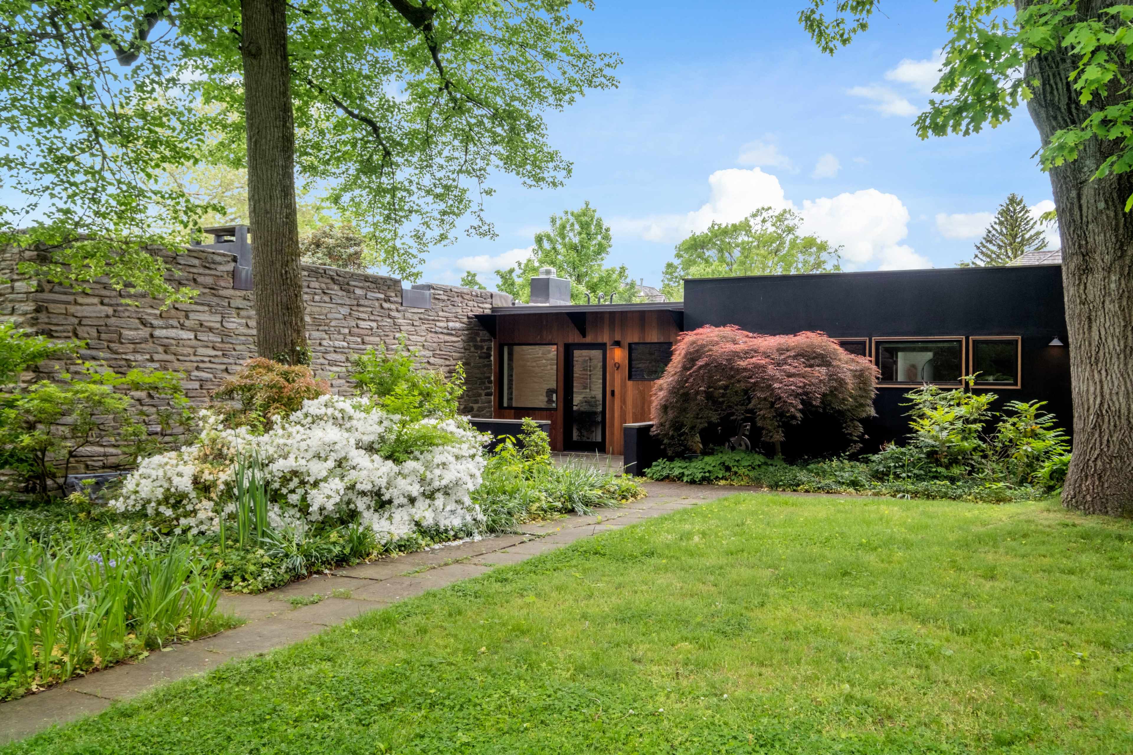 A modern house with a stone wall and landscaped garden featuring various plants and flowers lines a stone pathway.
