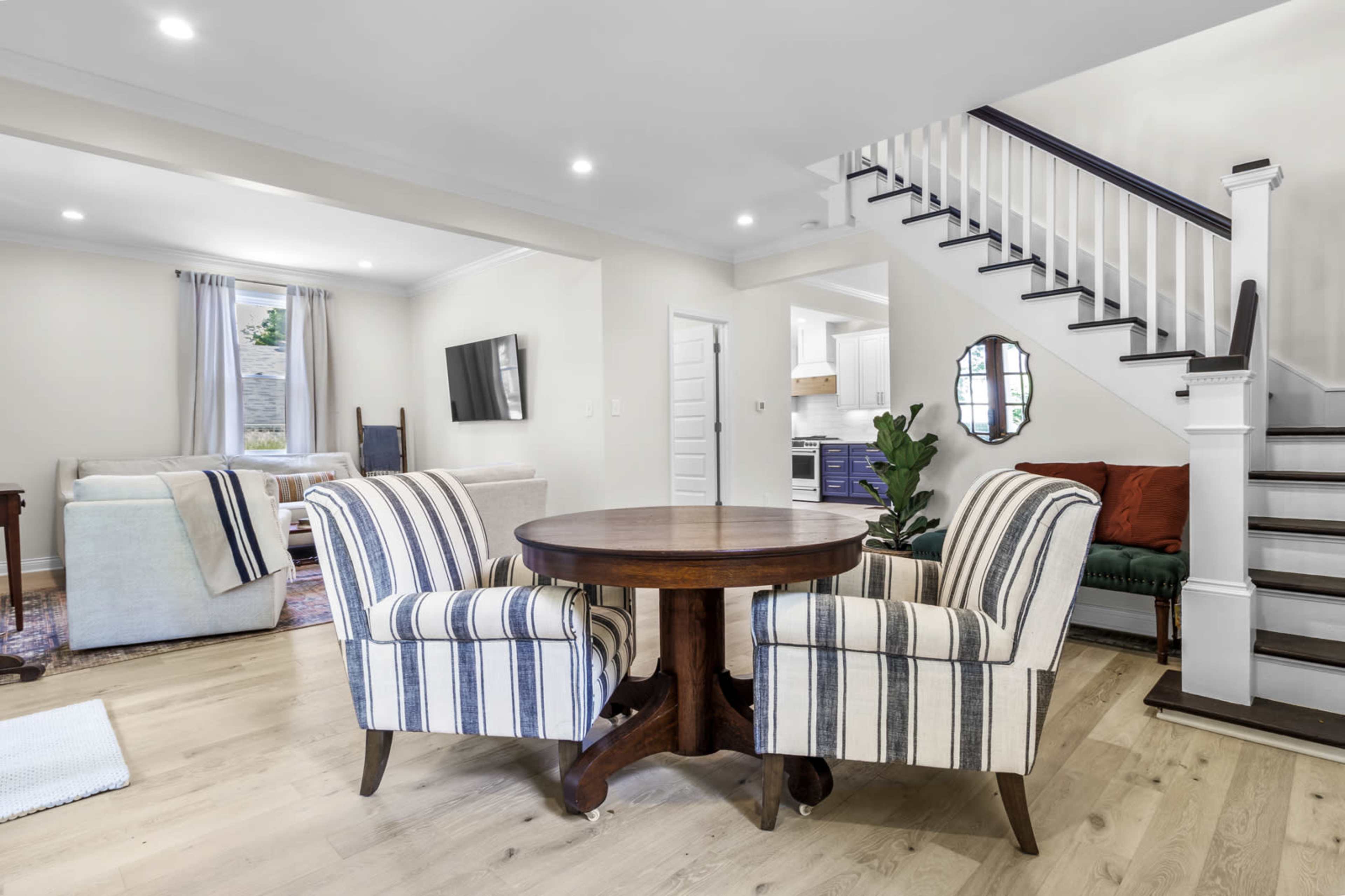 A modern interior featuring a round wooden dining table with two striped chairs, a staircase, and a living area visible in the background.