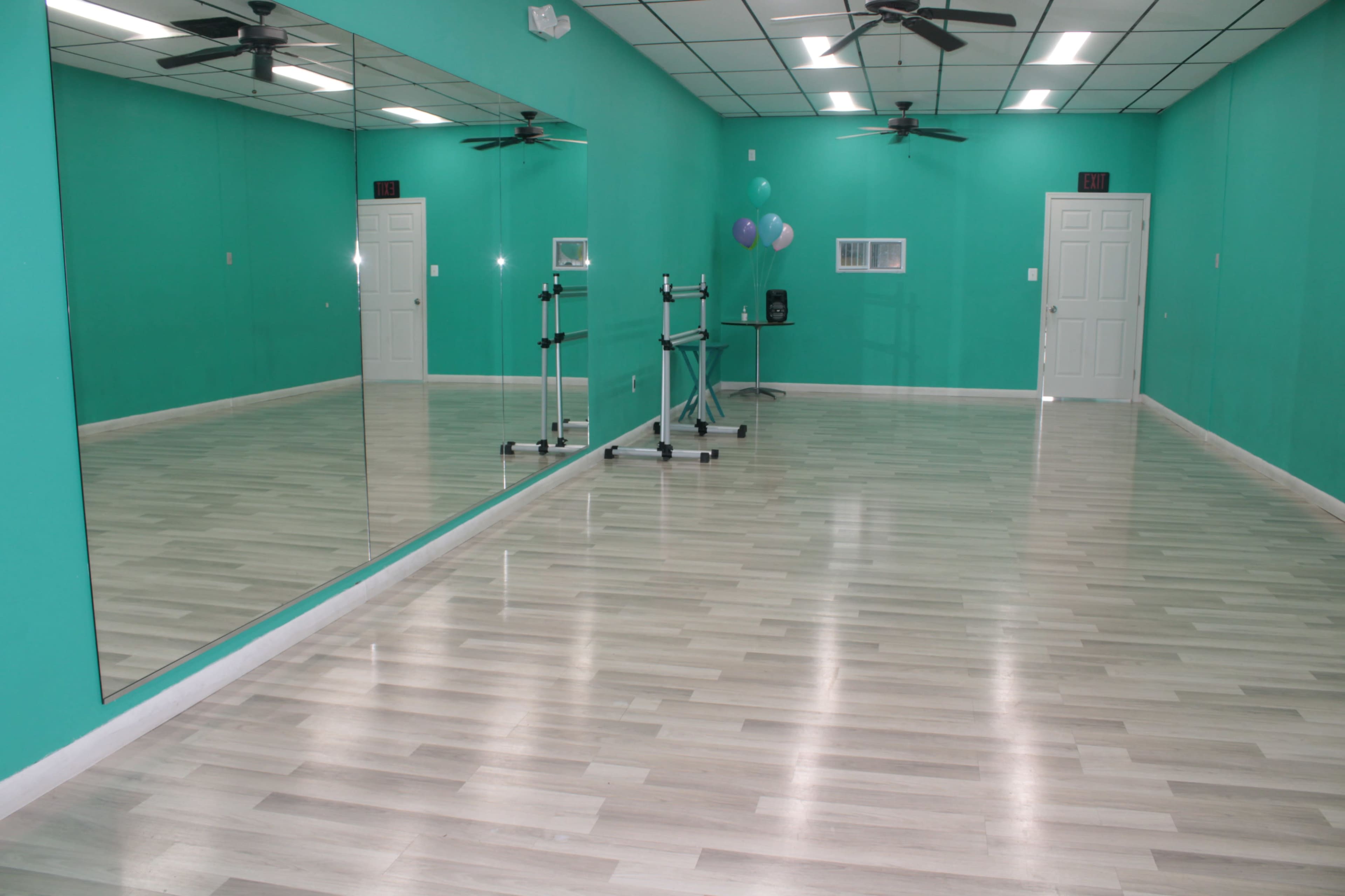 A bright, empty dance studio with teal walls, wooden floors, and large mirrors reflecting the space.