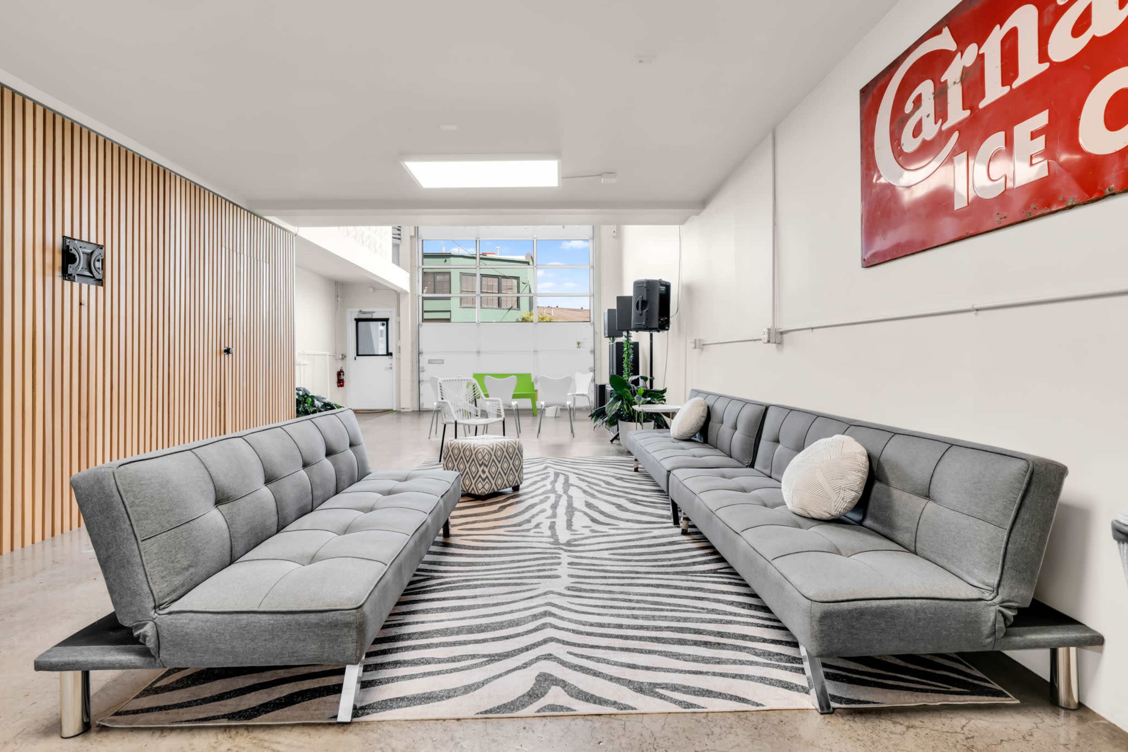 The image shows a modern interior space with two gray sofas aligned facing each other on a zebra-pattern rug, featuring large windows that allow natural light to enter.