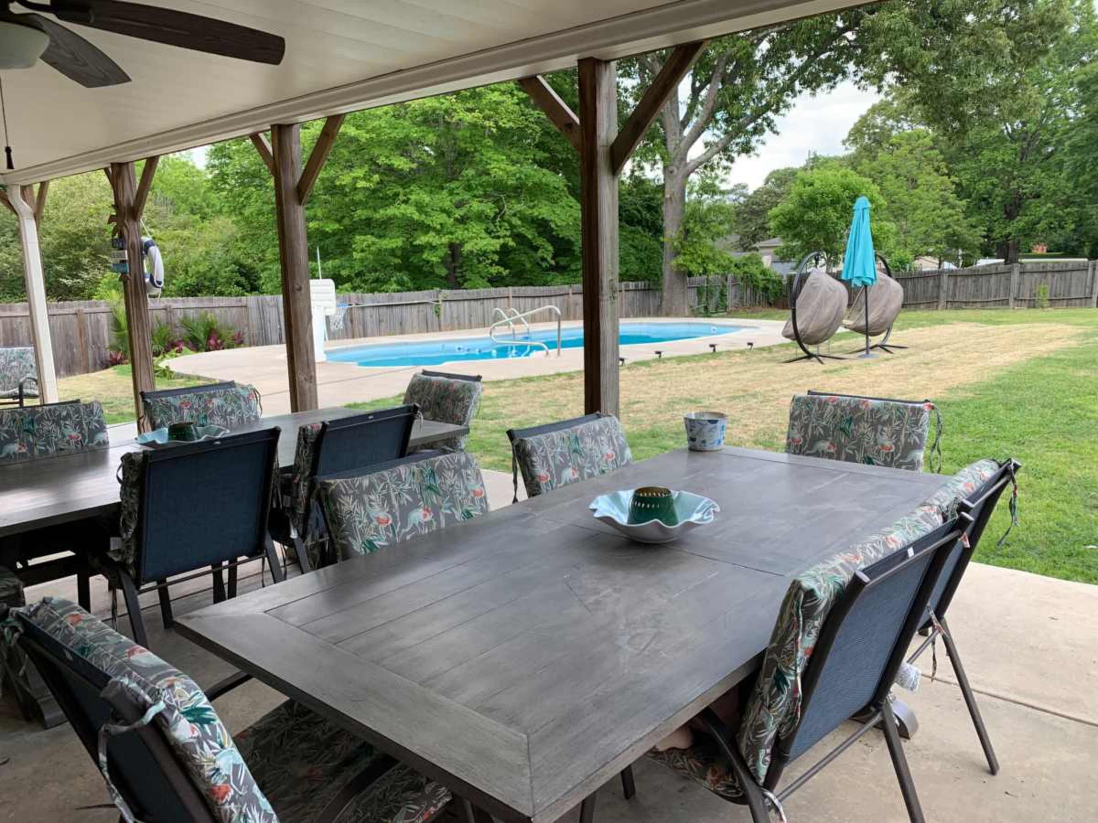 A covered patio with a large table and chairs overlooks a swimming pool and well-maintained yard.