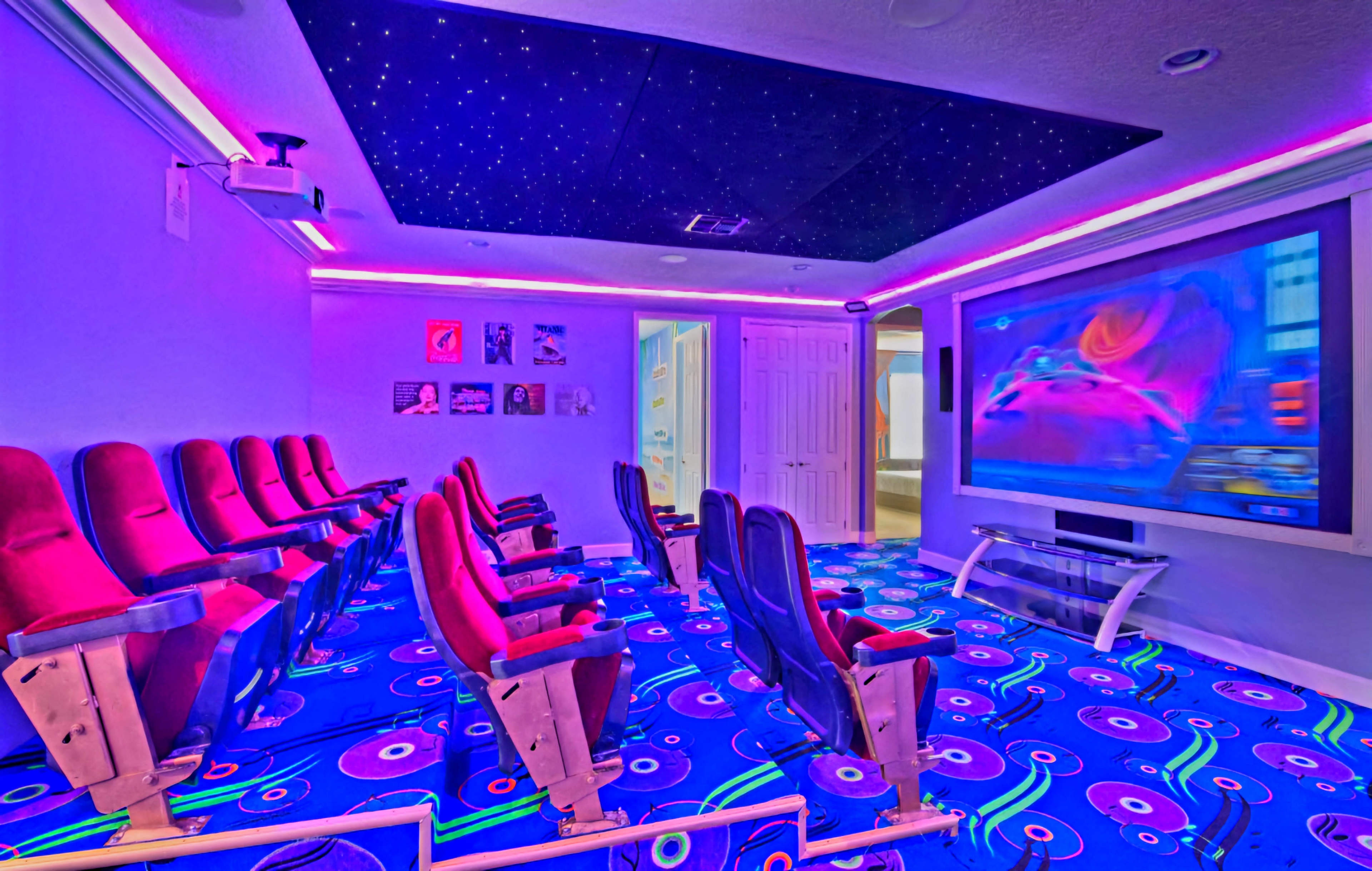 A home theater features red and black seating arranged in front of a large screen, with colorful carpet and a starry ceiling.