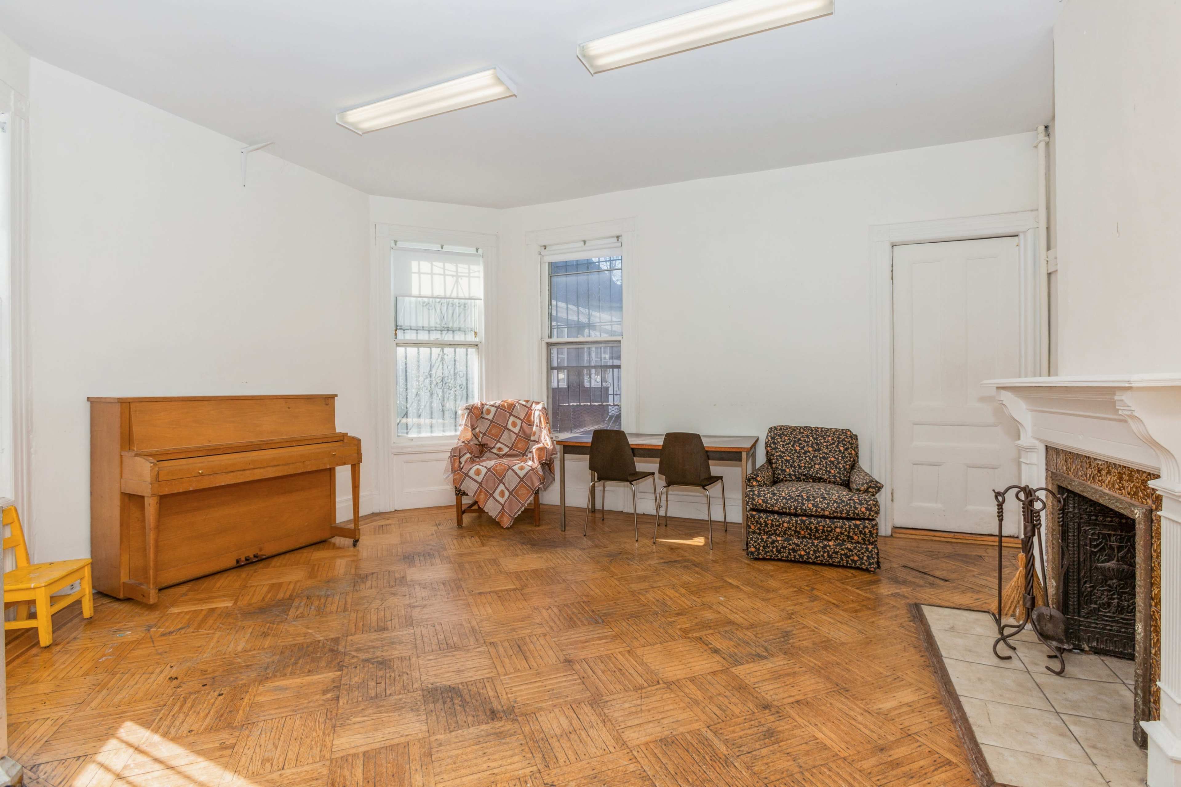 The image shows a sparse room with a piano, two chairs, and a patterned armchair surrounded by large windows and a fireplace.