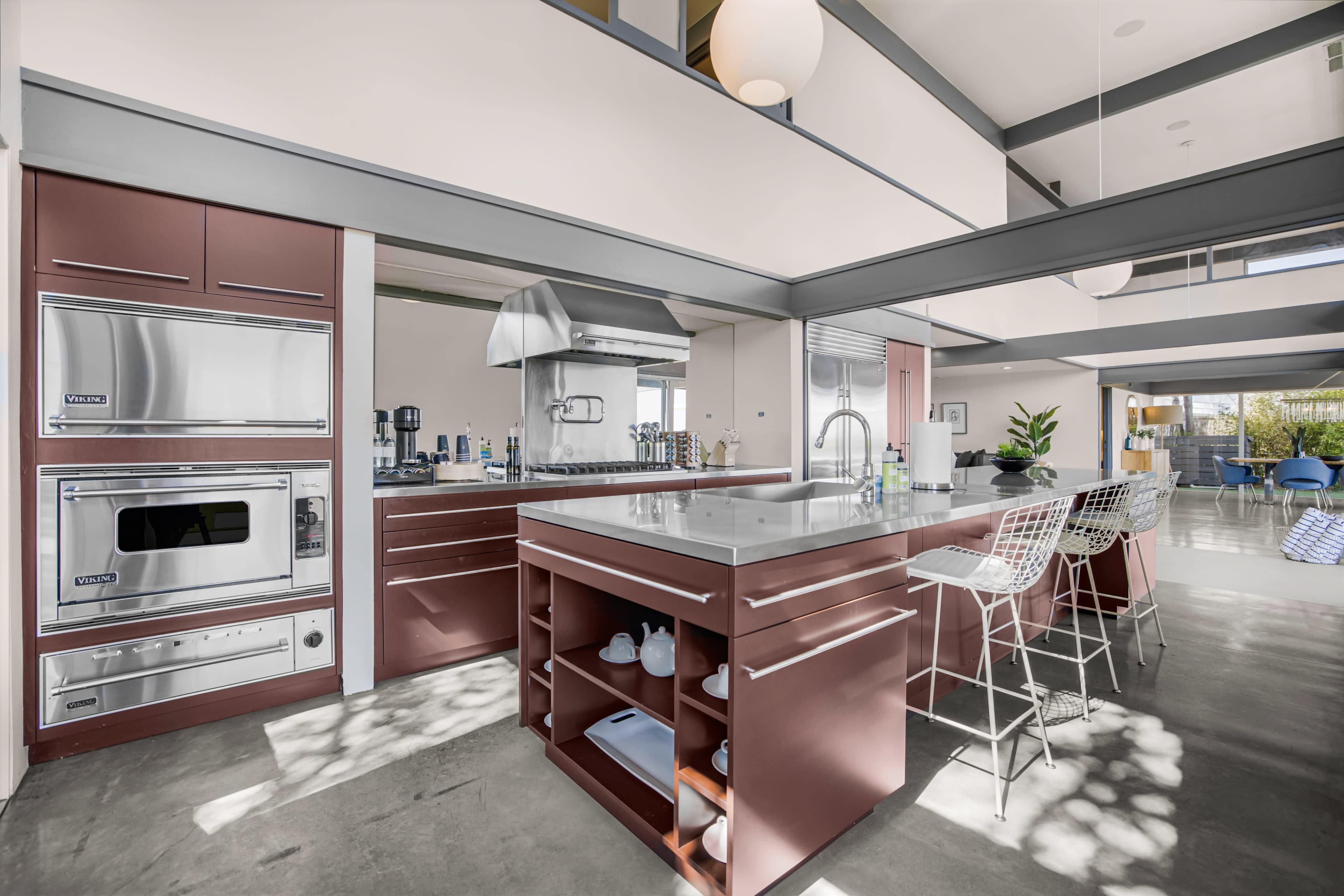 The image shows a modern kitchen with a central island, stainless steel appliances, and bar stools, featuring large windows that provide natural light.