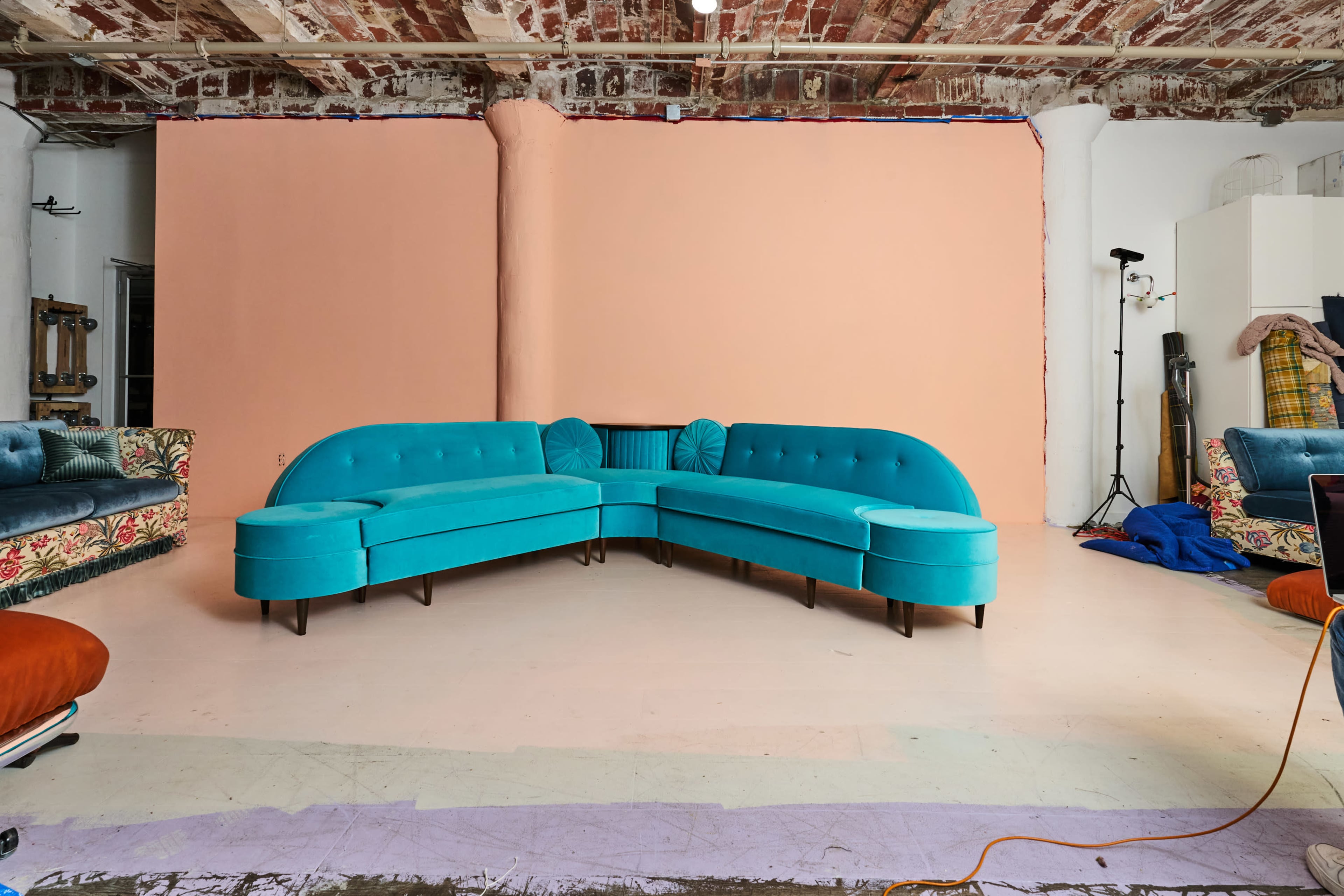 The image shows a turquoise curved sectional sofa positioned in front of a peach-colored wall, with additional sofas and studio equipment visible in the background.