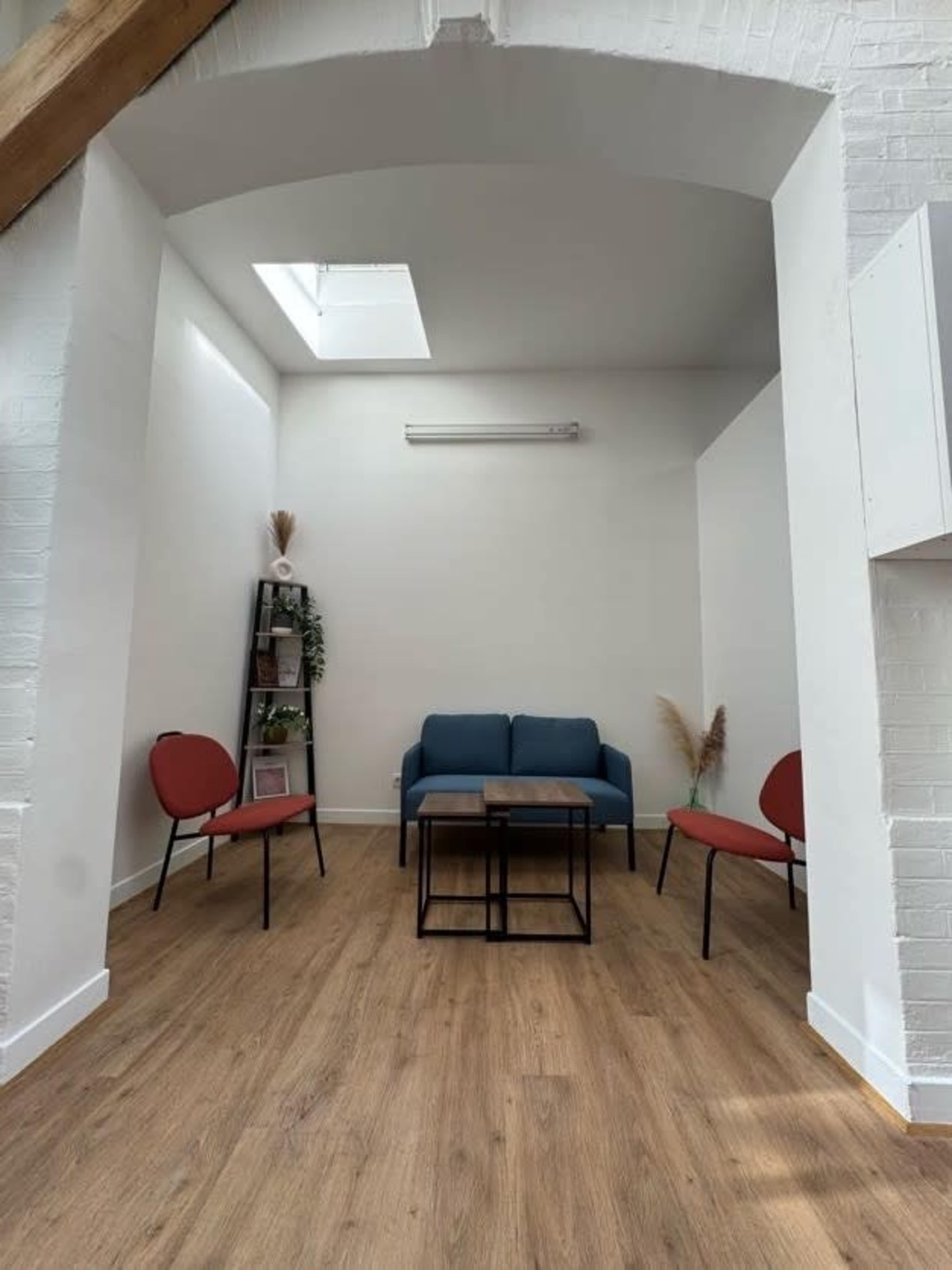The image shows a minimalist room featuring a blue couch, a coffee table, and four chairs arranged around a wooden floor with a skylight above.