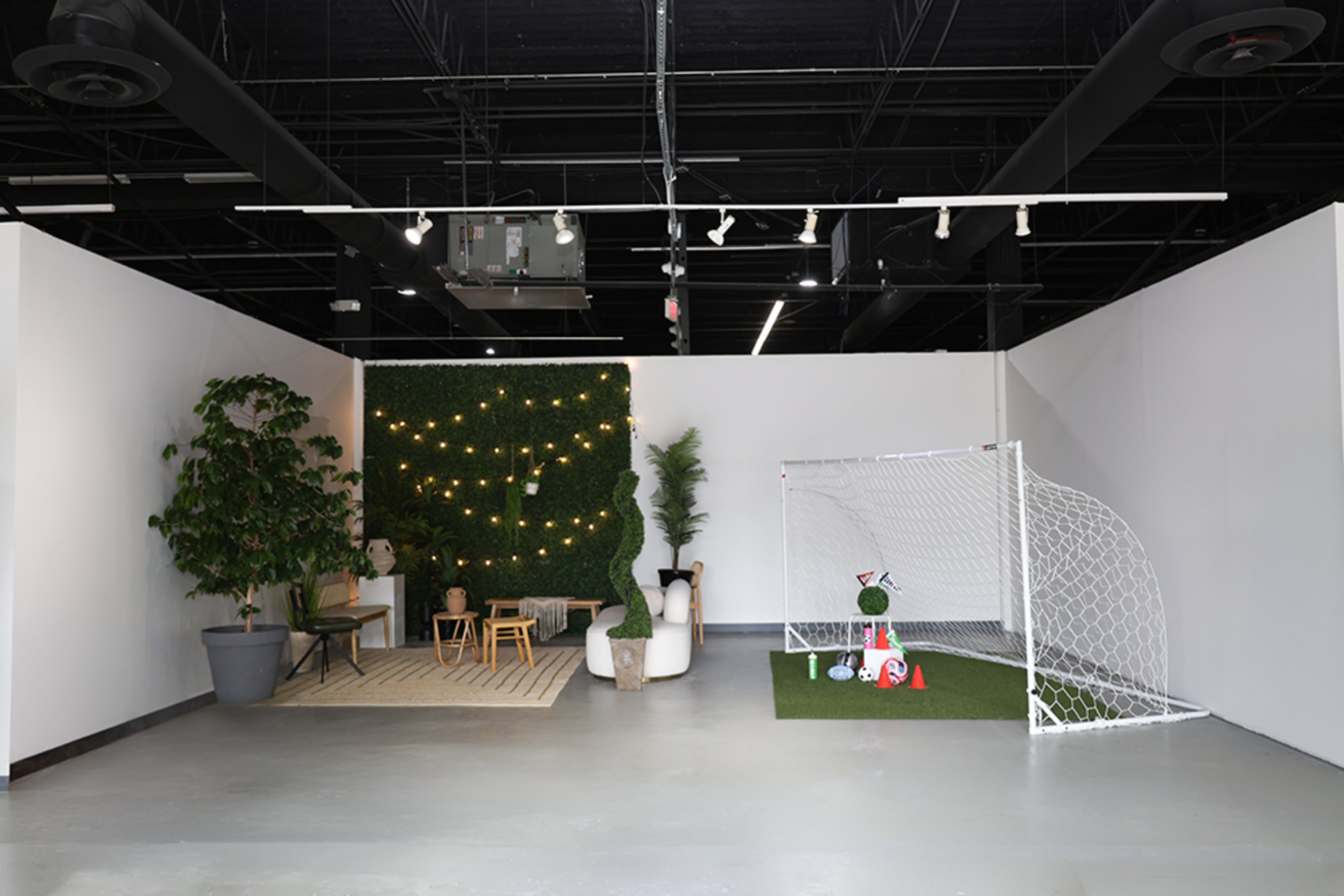 The image depicts an indoor space with a small soccer goal on artificial grass, a green wall with lights, and seating arrangements including a small table and chairs.