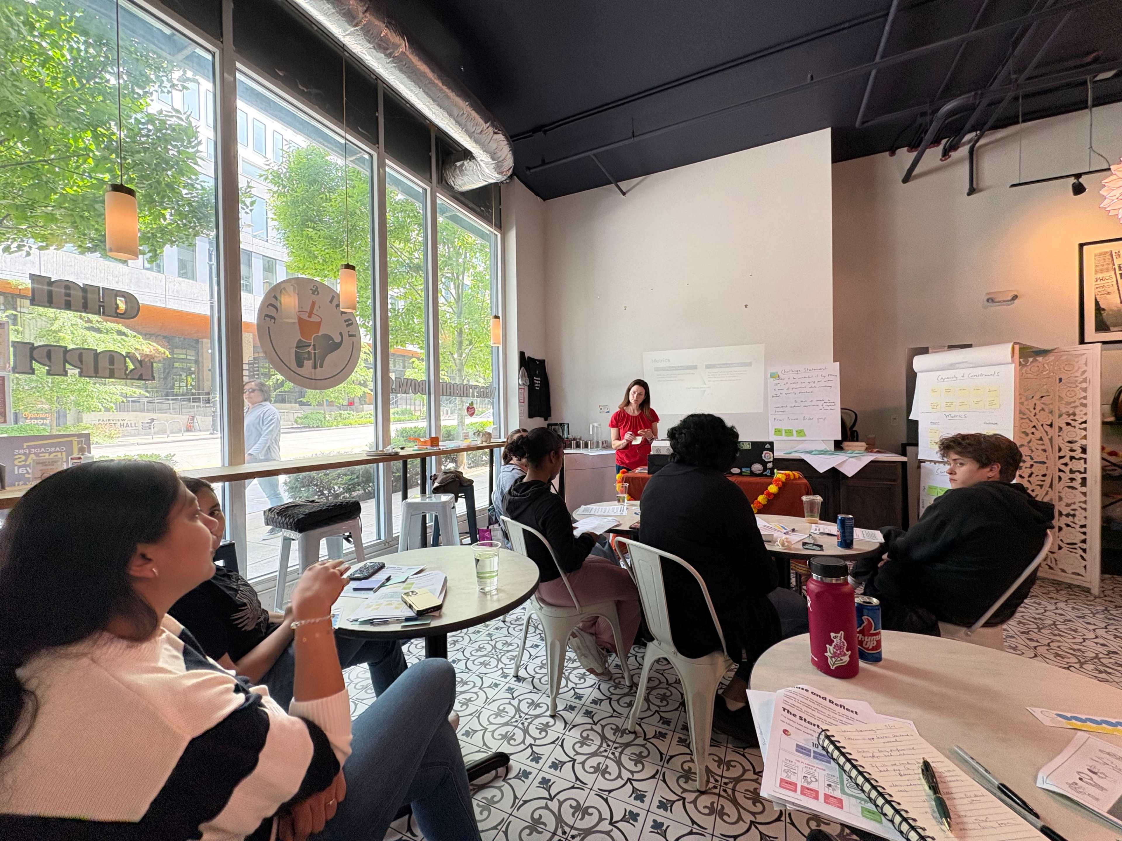 A group of people sits at tables in a café while a woman stands and presents at the front of the room.