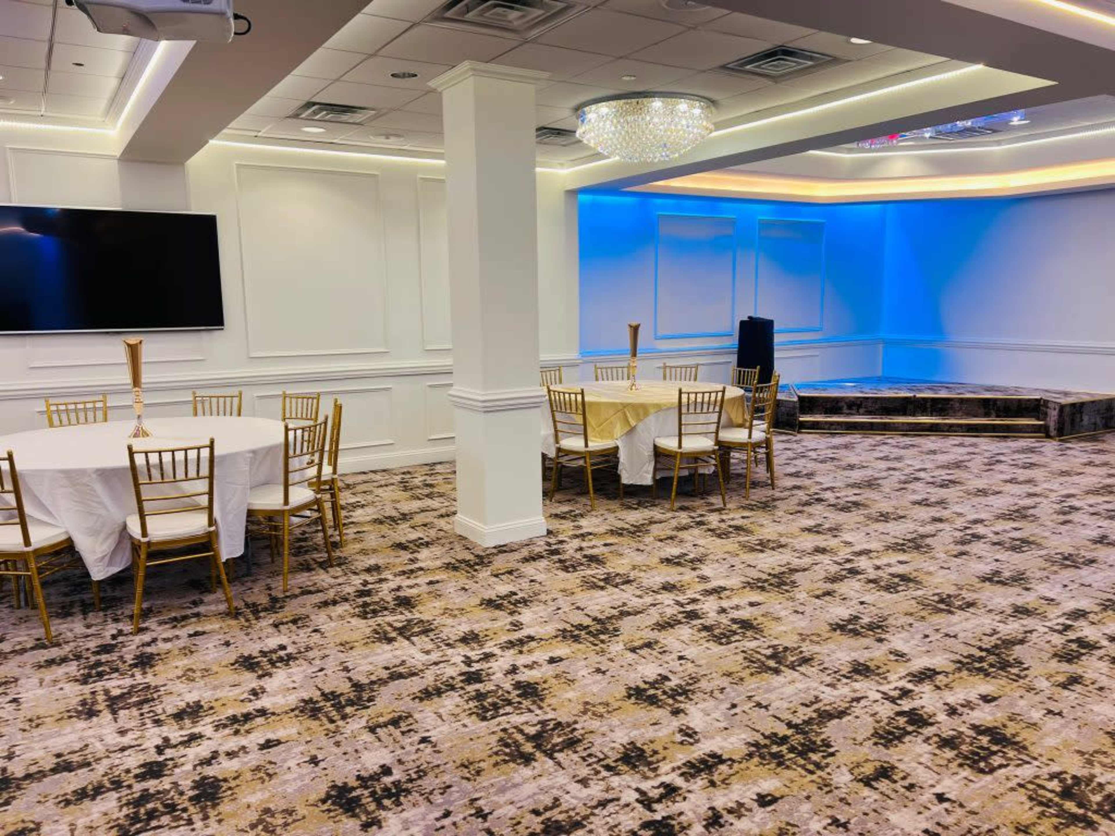 The image shows a spacious banquet room with several round tables set for dining, a stage area, and soft lighting highlights on the walls.