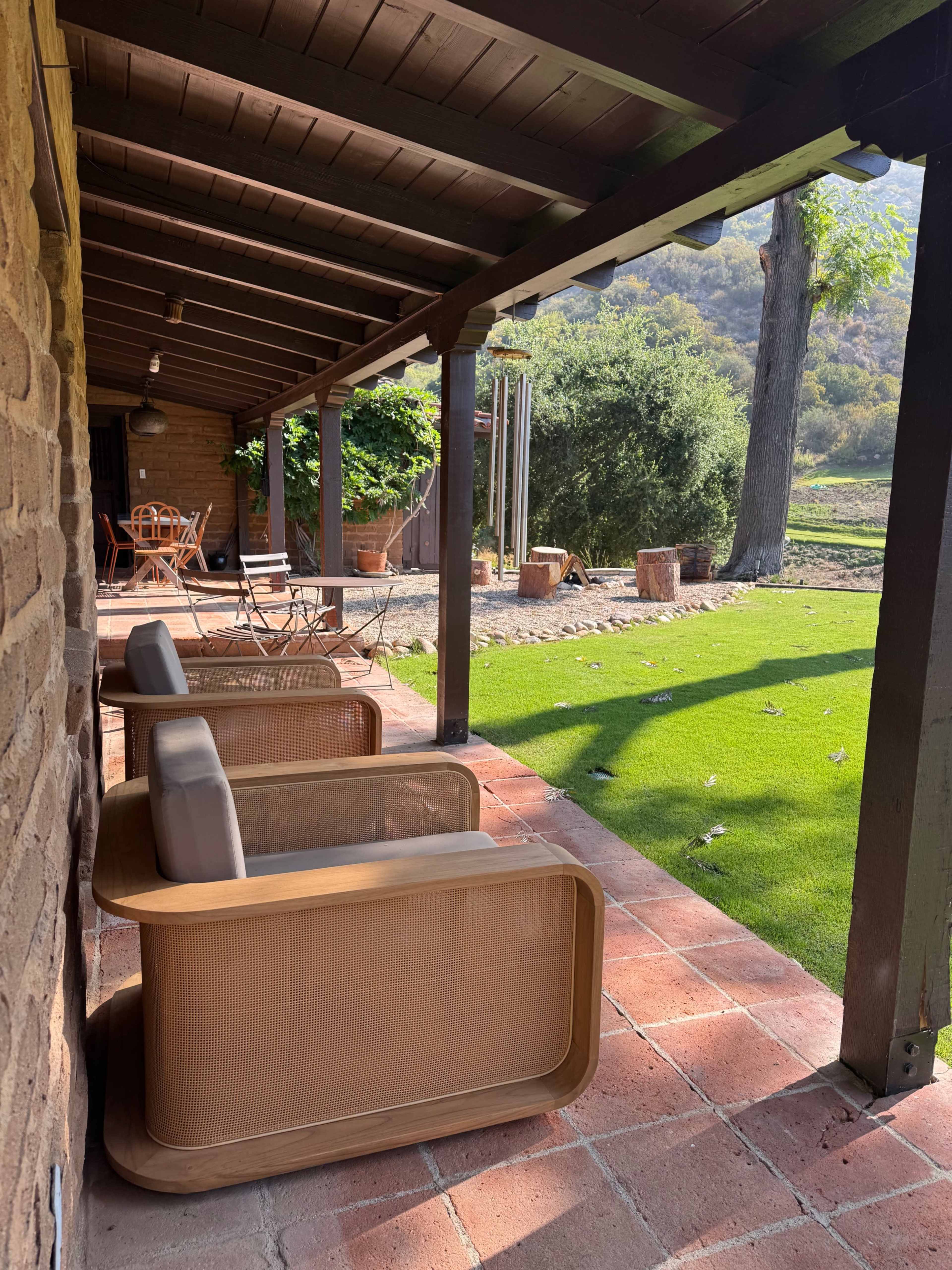 A porch features three modern chairs arranged along a tiled pathway, overlooking a green lawn and trees.
