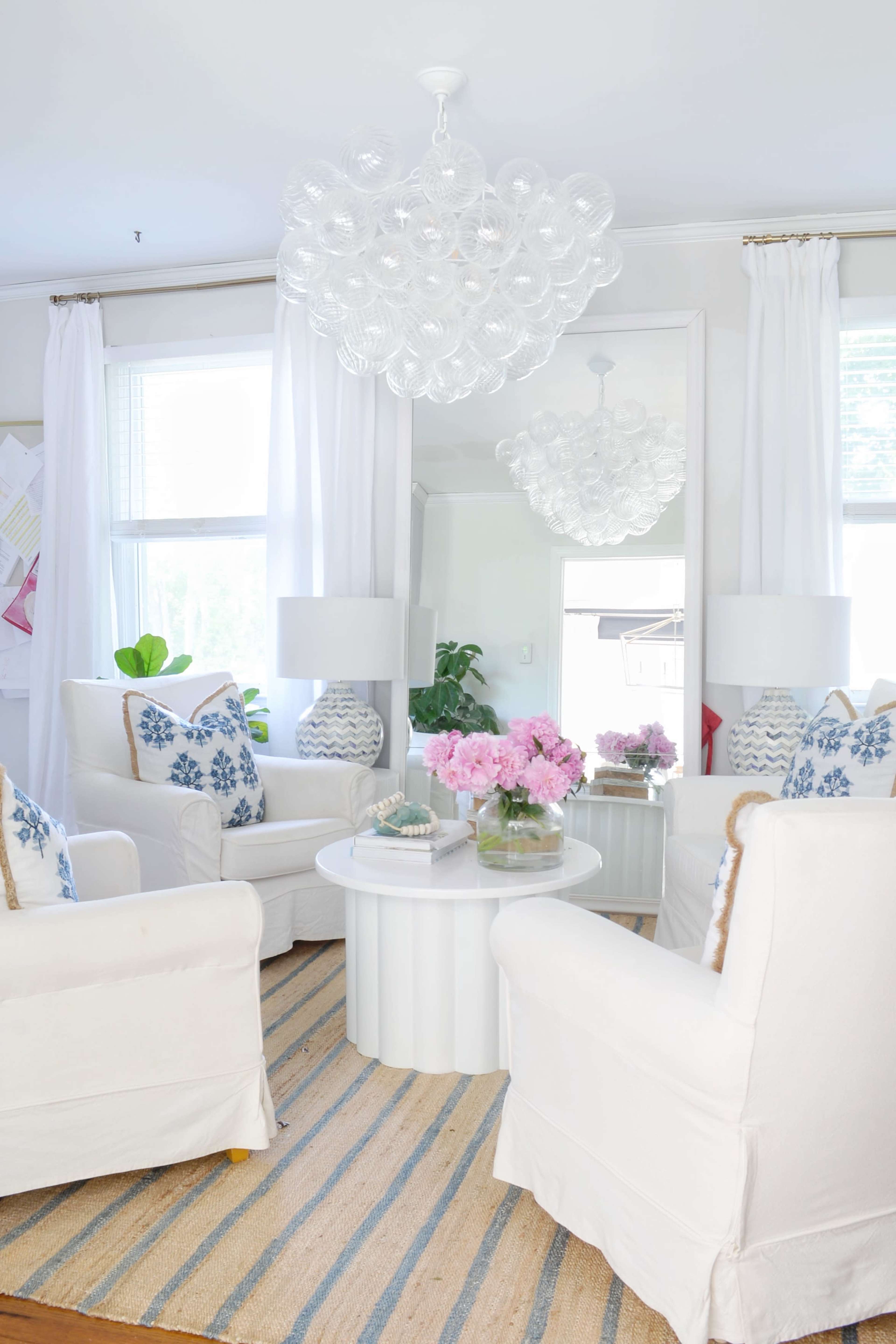 A bright living room with white upholstered chairs, a round coffee table, and a large chandelier, featuring floral pillows and pink flowers in vases.