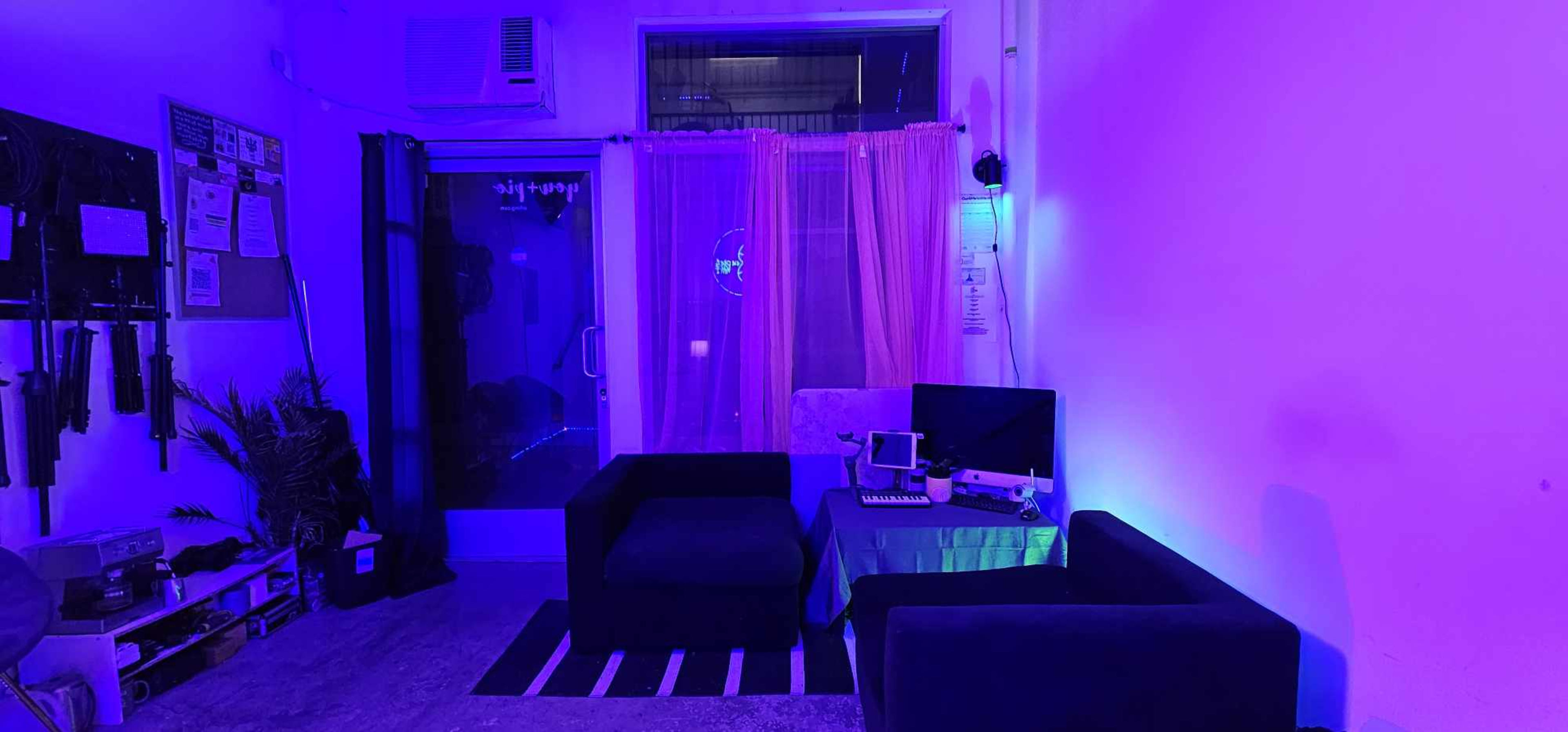 The image shows a small room with purple ambient lighting, featuring two dark sofas, a table with equipment, and a doorway with curtains.