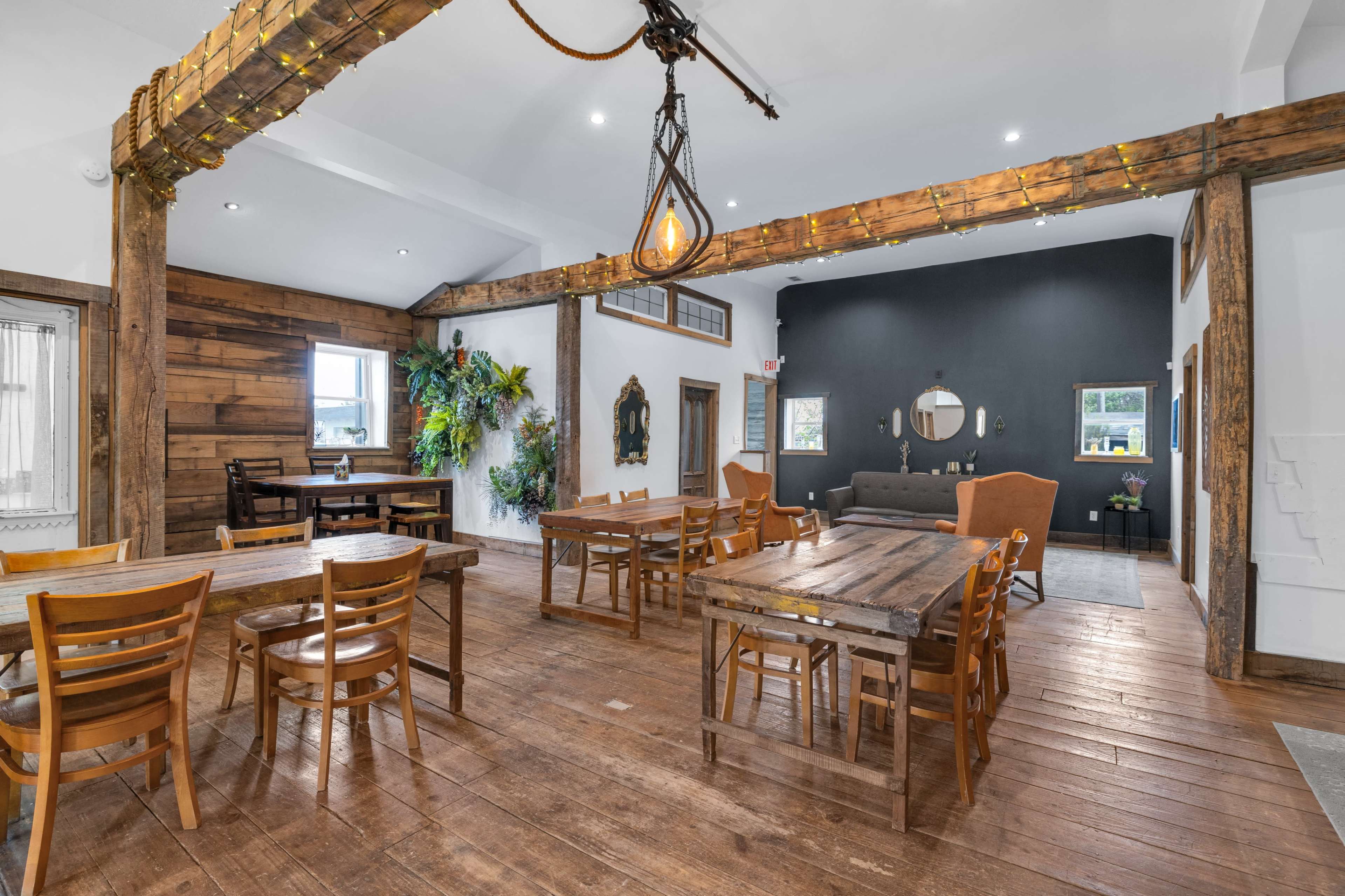 The image shows a rustic interior space with wooden tables, chairs, and a mix of decor, including plants and artwork, in a well-lit room featuring exposed beams and a contrasting dark wall.