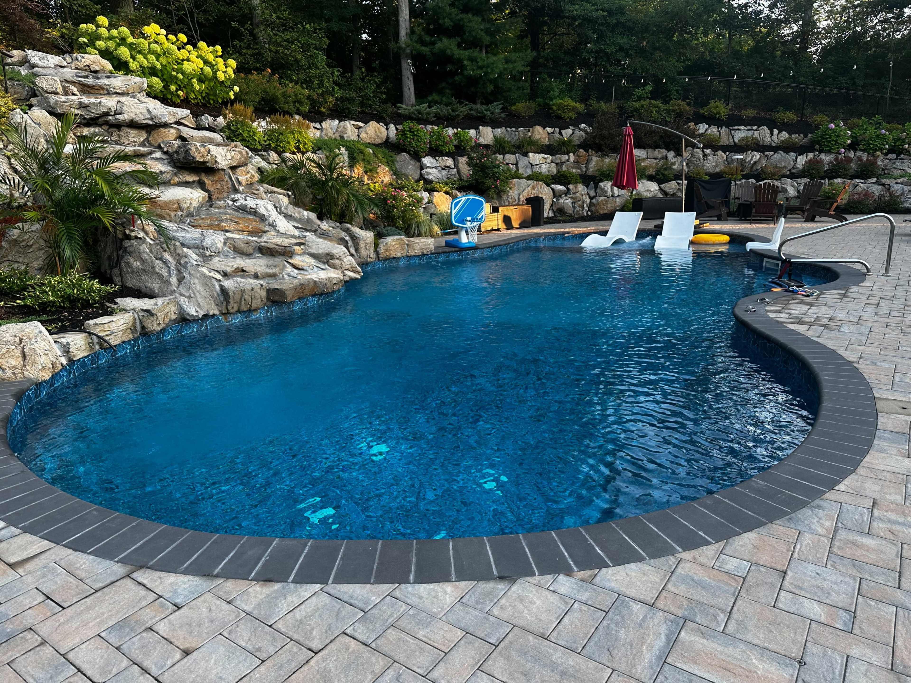The image shows a blue swimming pool surrounded by rock landscaping and potted plants.