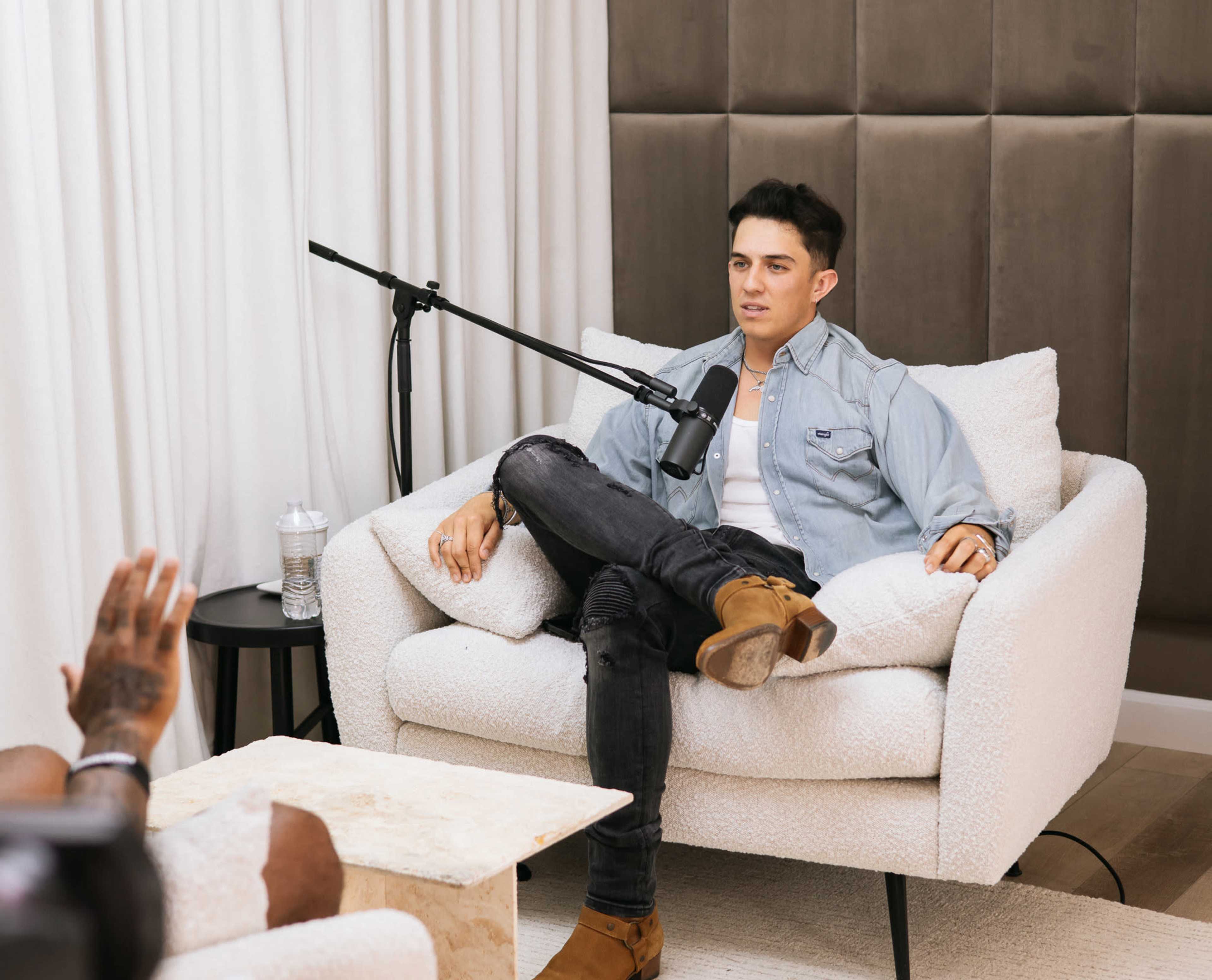 A man sits in a light-colored armchair with a microphone positioned in front of him, engaged in conversation.