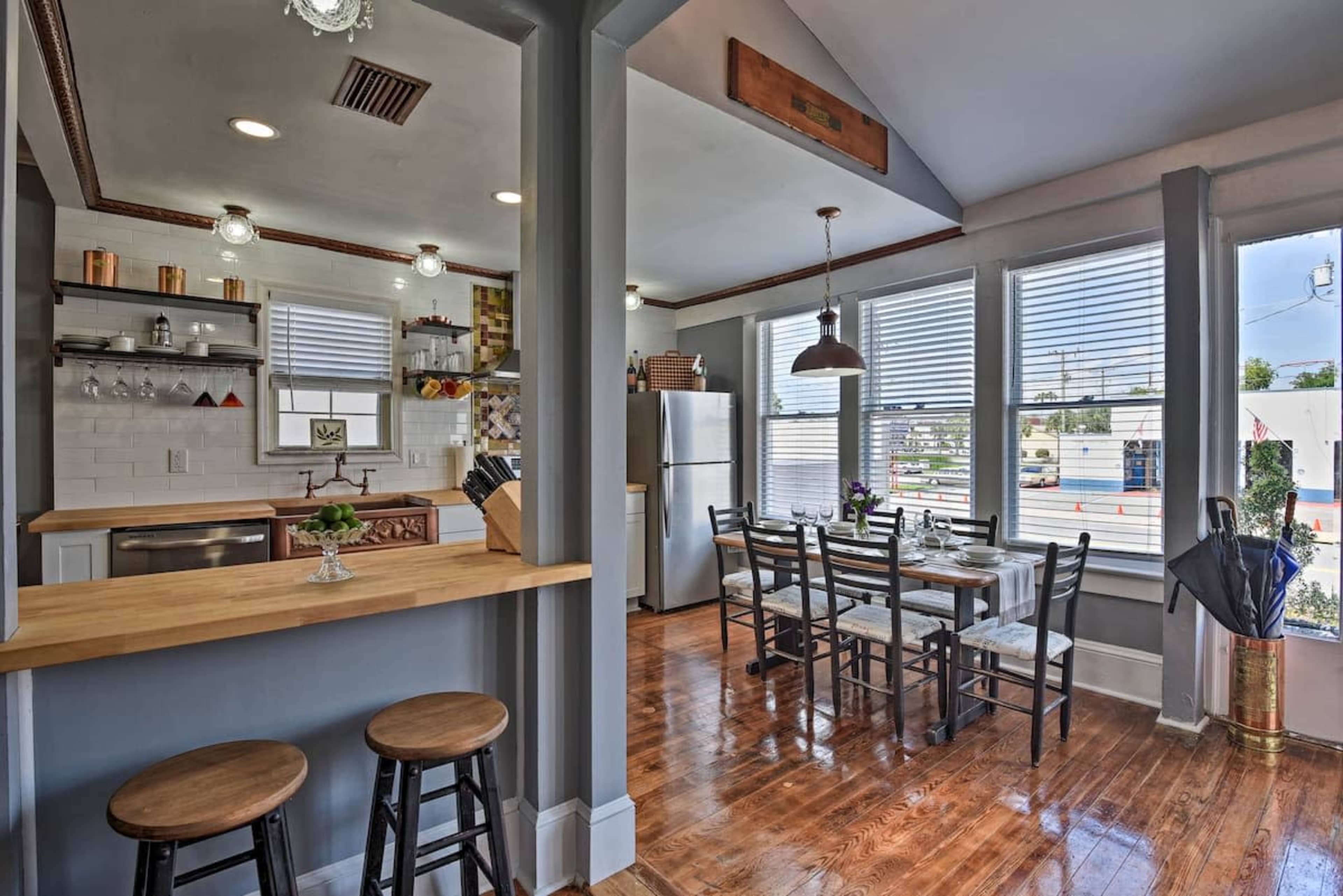 A kitchen and dining area feature wooden flooring, a bar counter with two stools, and a dining table set for four beneath large windows.