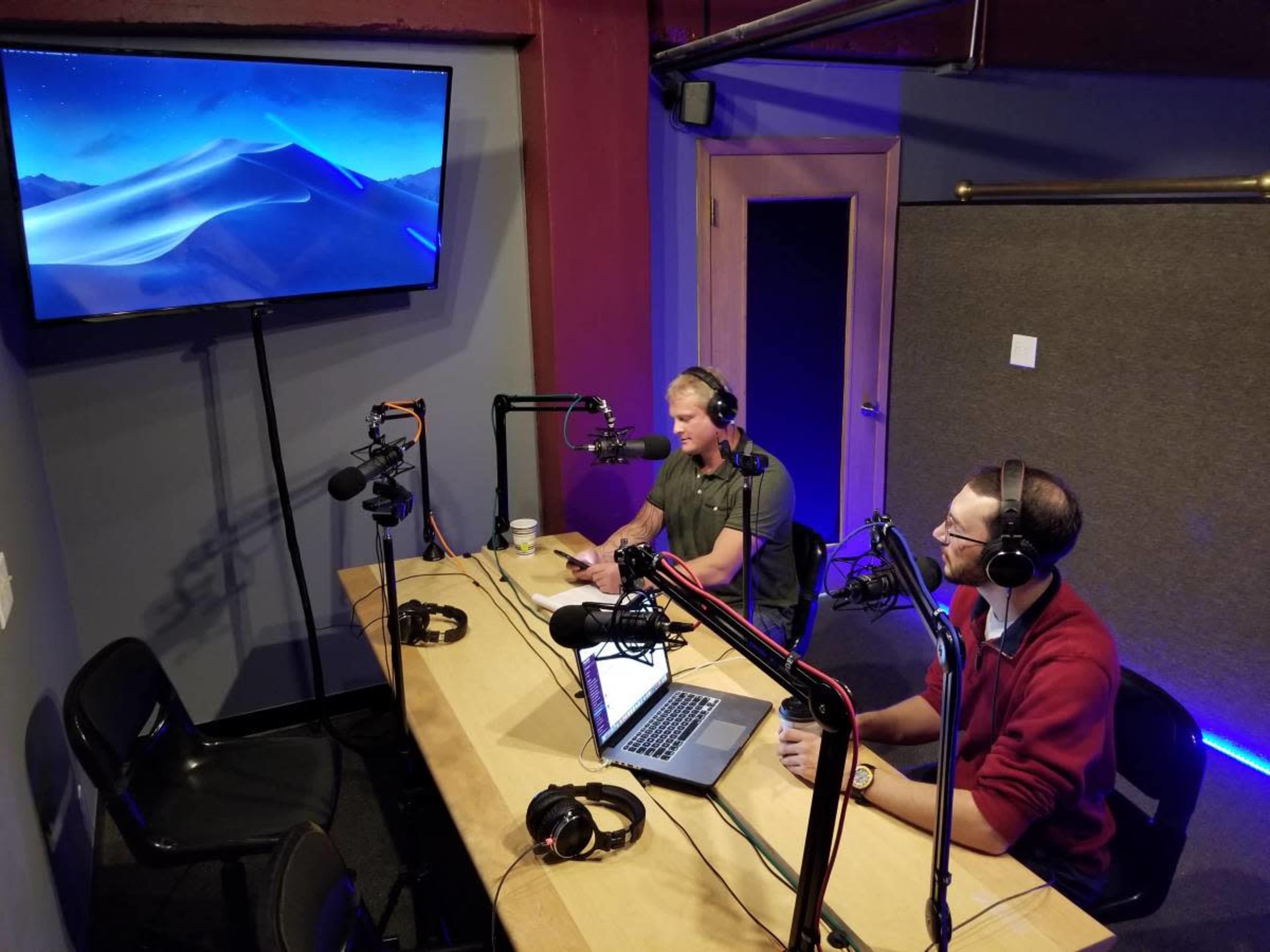 Two men are seated at a table in a recording studio, with microphones in front of them and a large screen displaying a mountain landscape in the background.