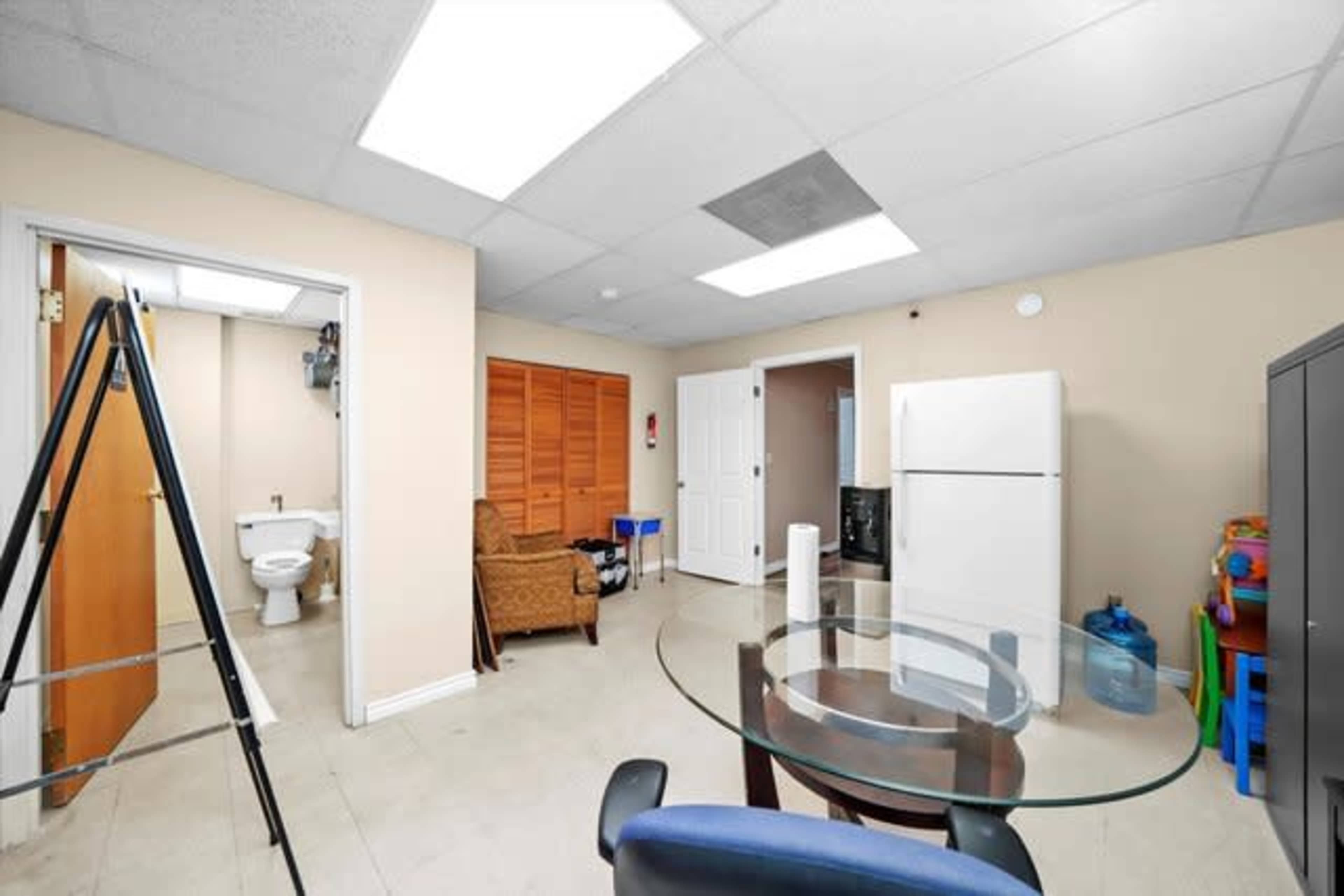 The image depicts a small room featuring a glass table, a chair, a refrigerator, a toilet, and a closet with wooden doors.