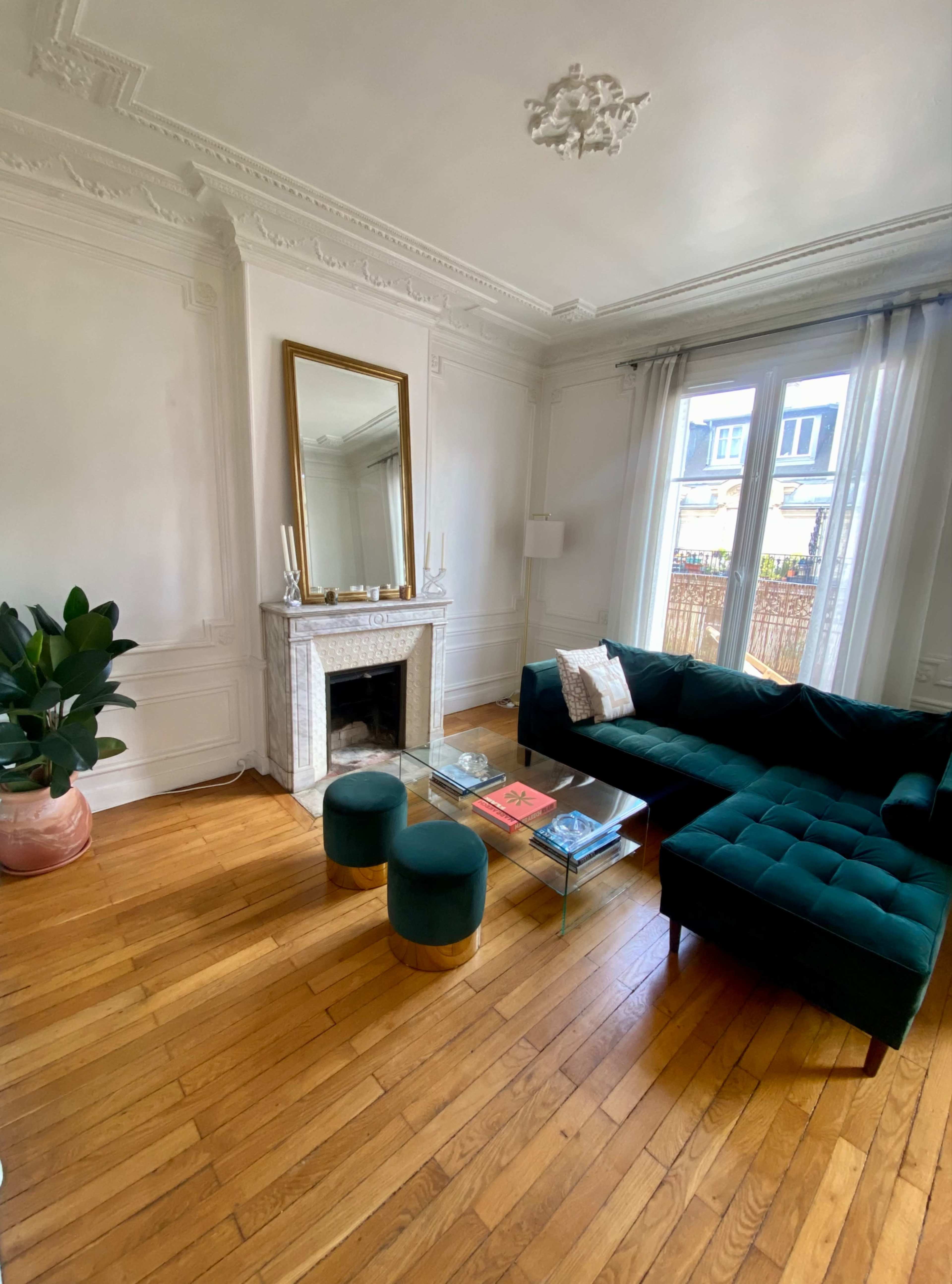 A bright living room with a green velvet sectional sofa, two round green ottomans, a marble fireplace, and a large mirror above it.
