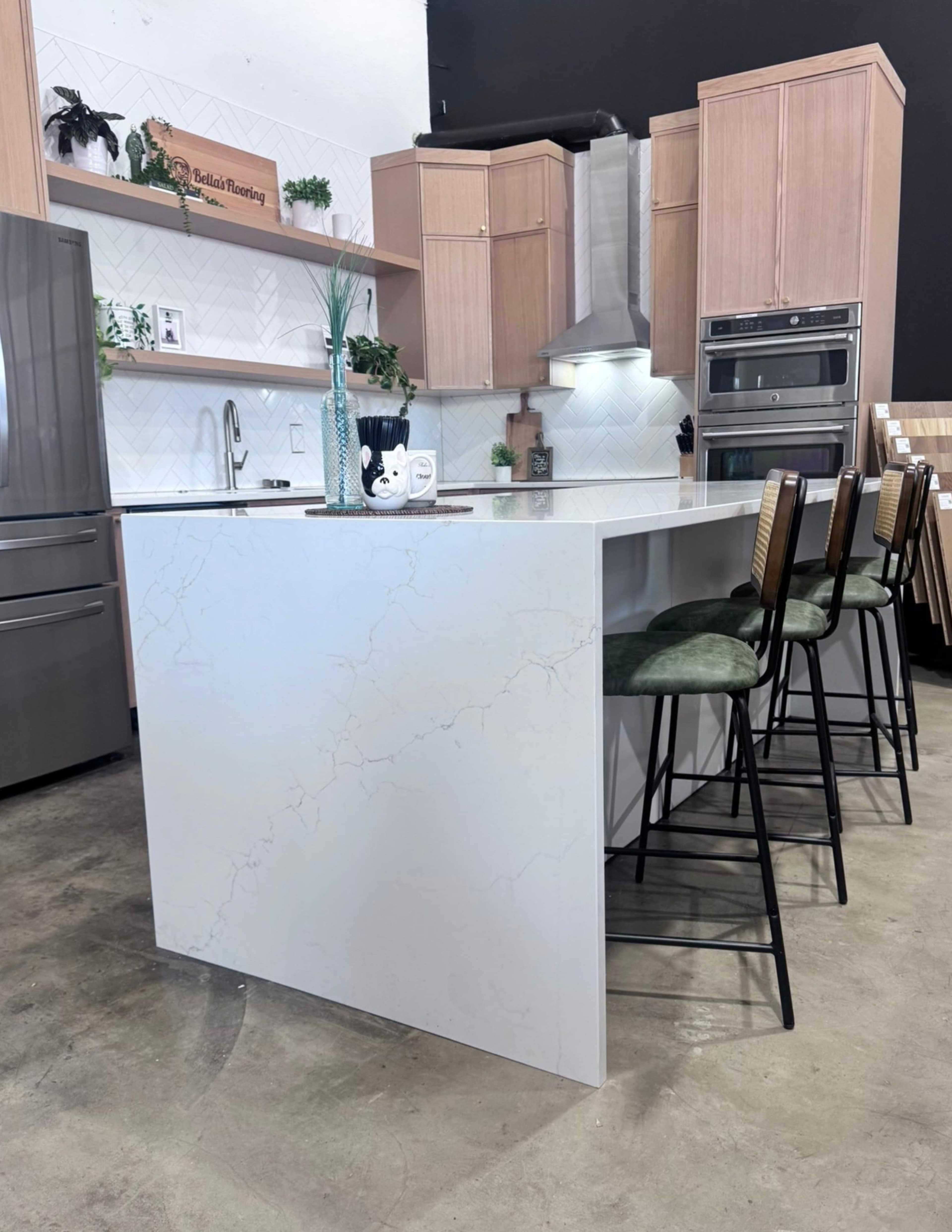 The image shows a modern kitchen with a large island featuring a white countertop, bar stools, wooden cabinetry, and stainless steel appliances.