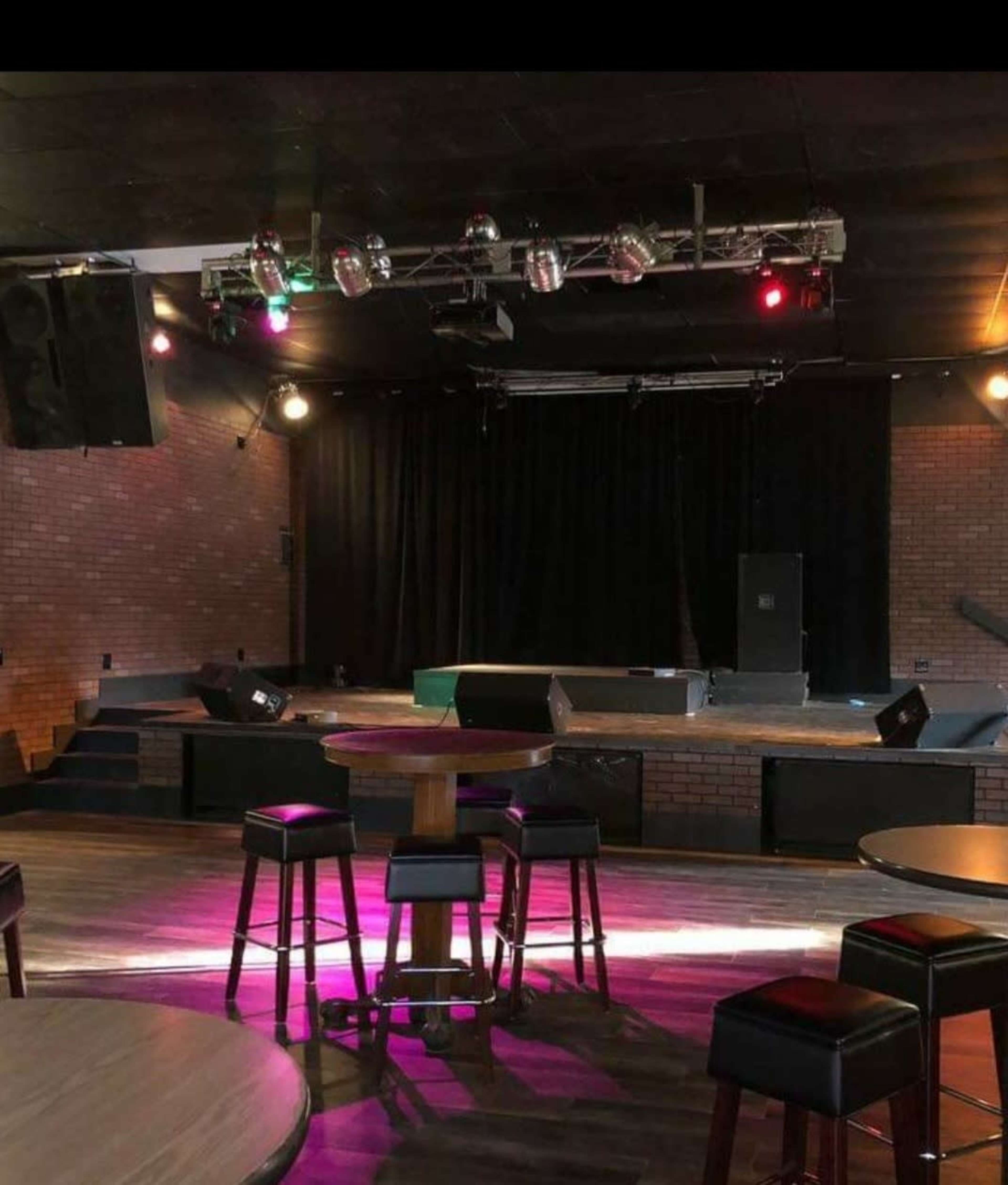 The image shows a music venue with a stage at the front, equipped with lights and seating arrangements of tables and stools.