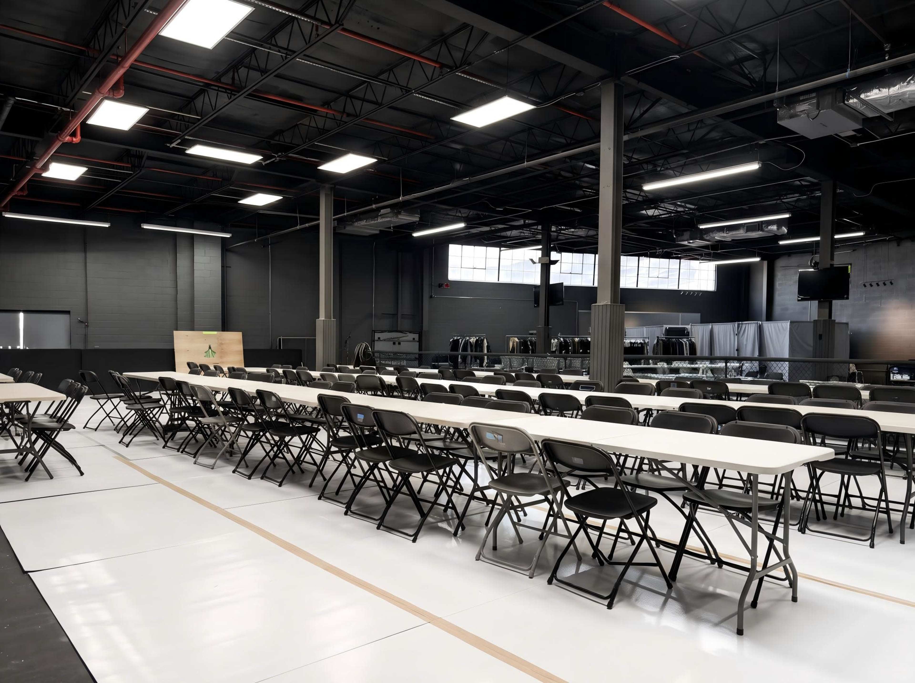 The image shows a spacious indoor area set up with rows of folding chairs and tables, illuminated by overhead lights.