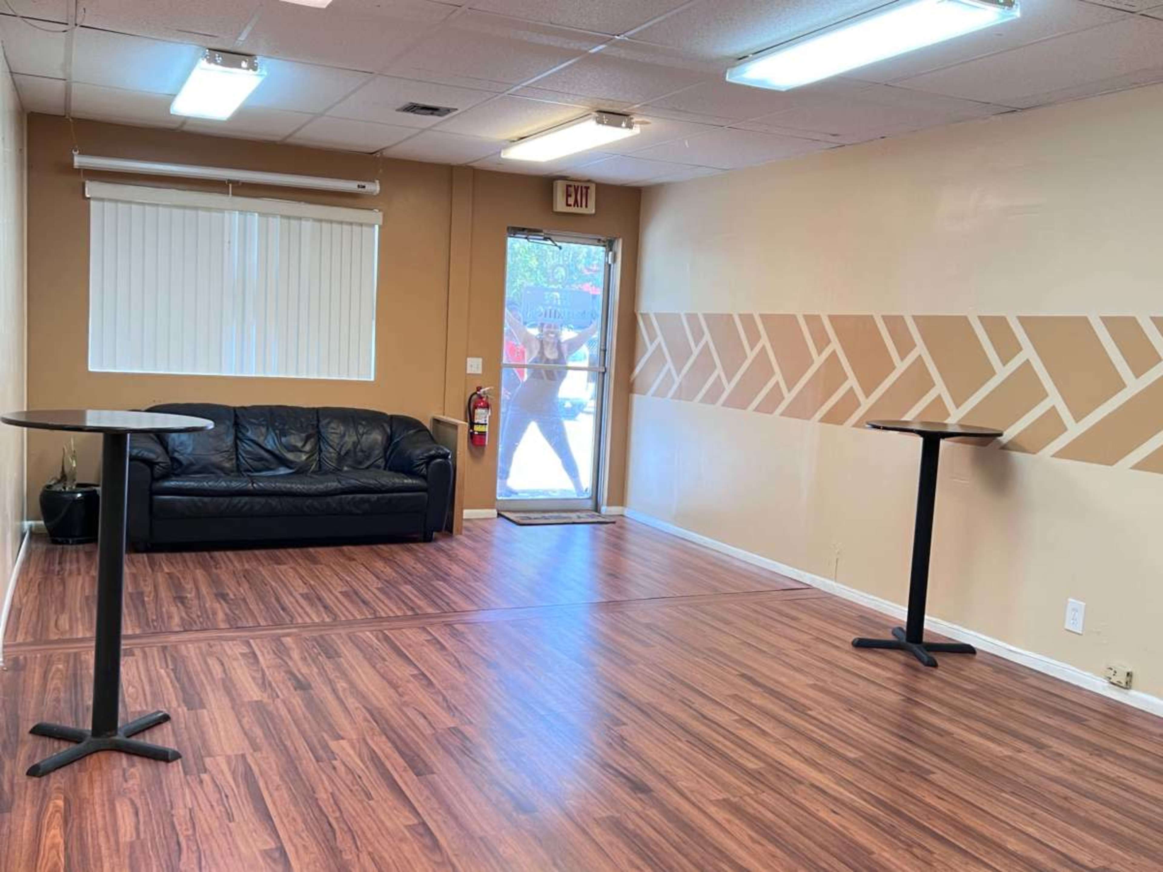 A spacious interior with a black couch, two small tables, and a door leading outside, all set on wooden flooring.