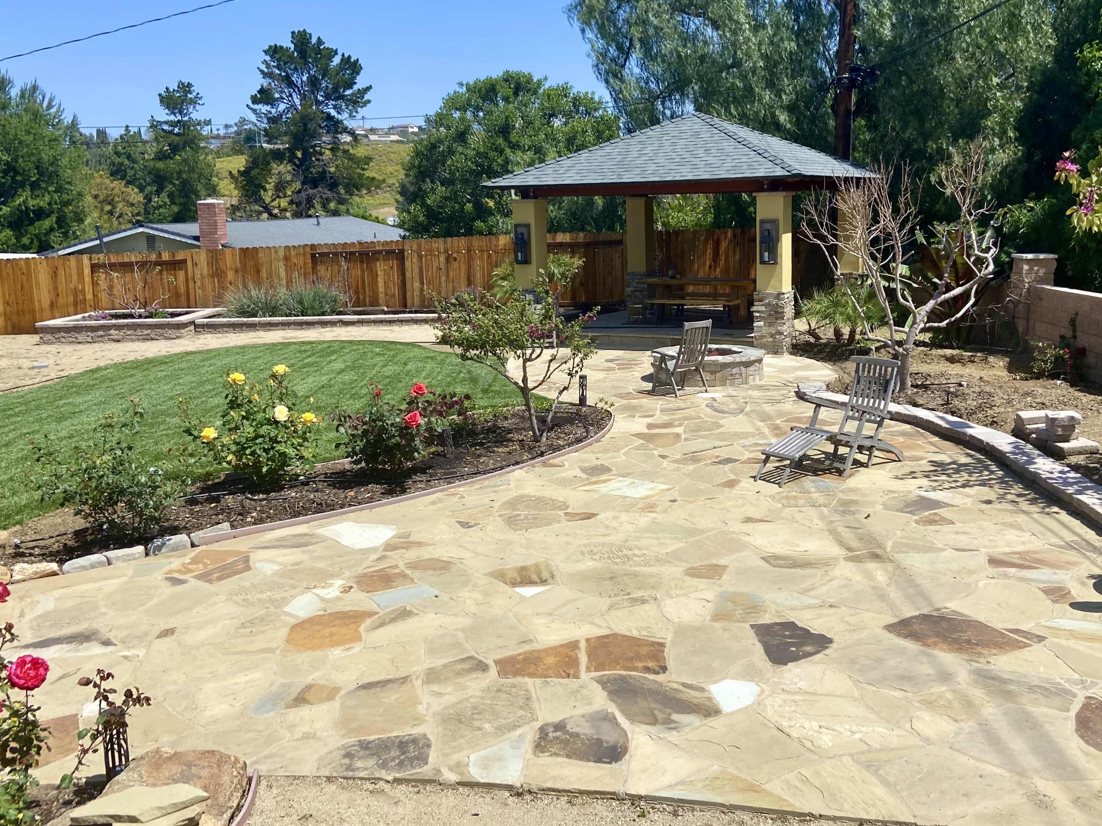 A landscaped backyard with a stone patio, a gazebo, flower beds with roses, and a grassy area surrounded by a wooden fence.